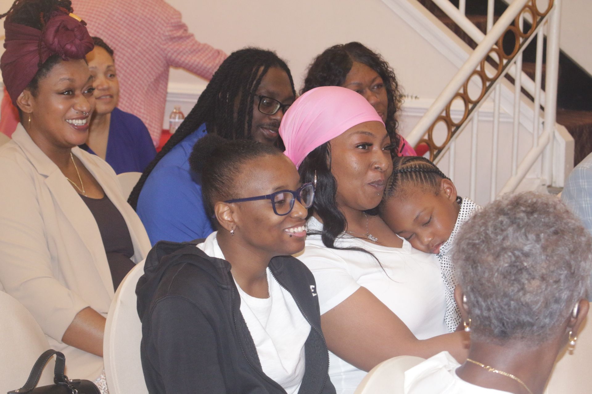 Audience seated indoors, with one woman holding a sleeping child in a pink headwrap and others smiling nearby