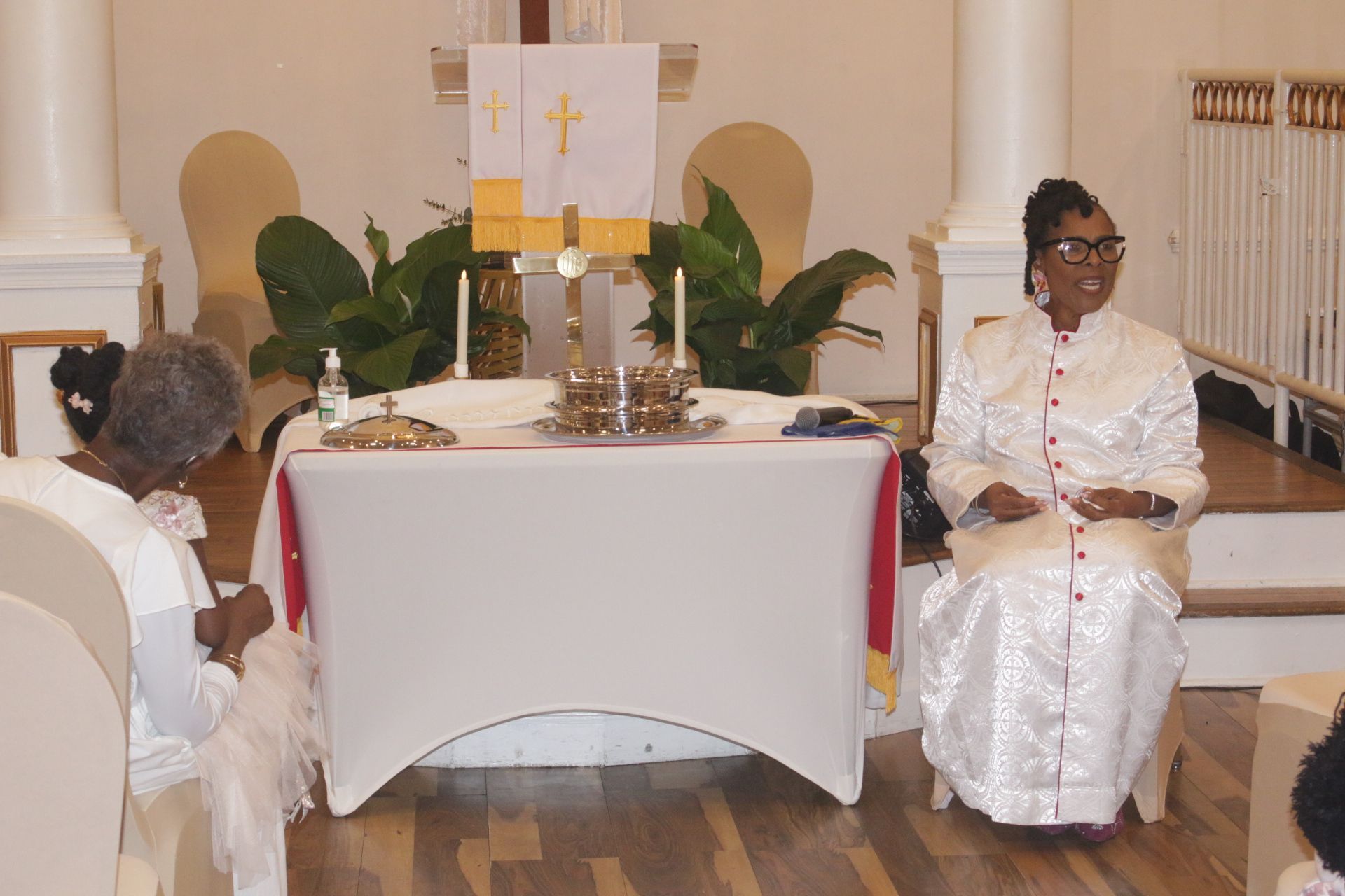 Two women in white sit beside a church altar with candles and a white cloth.
