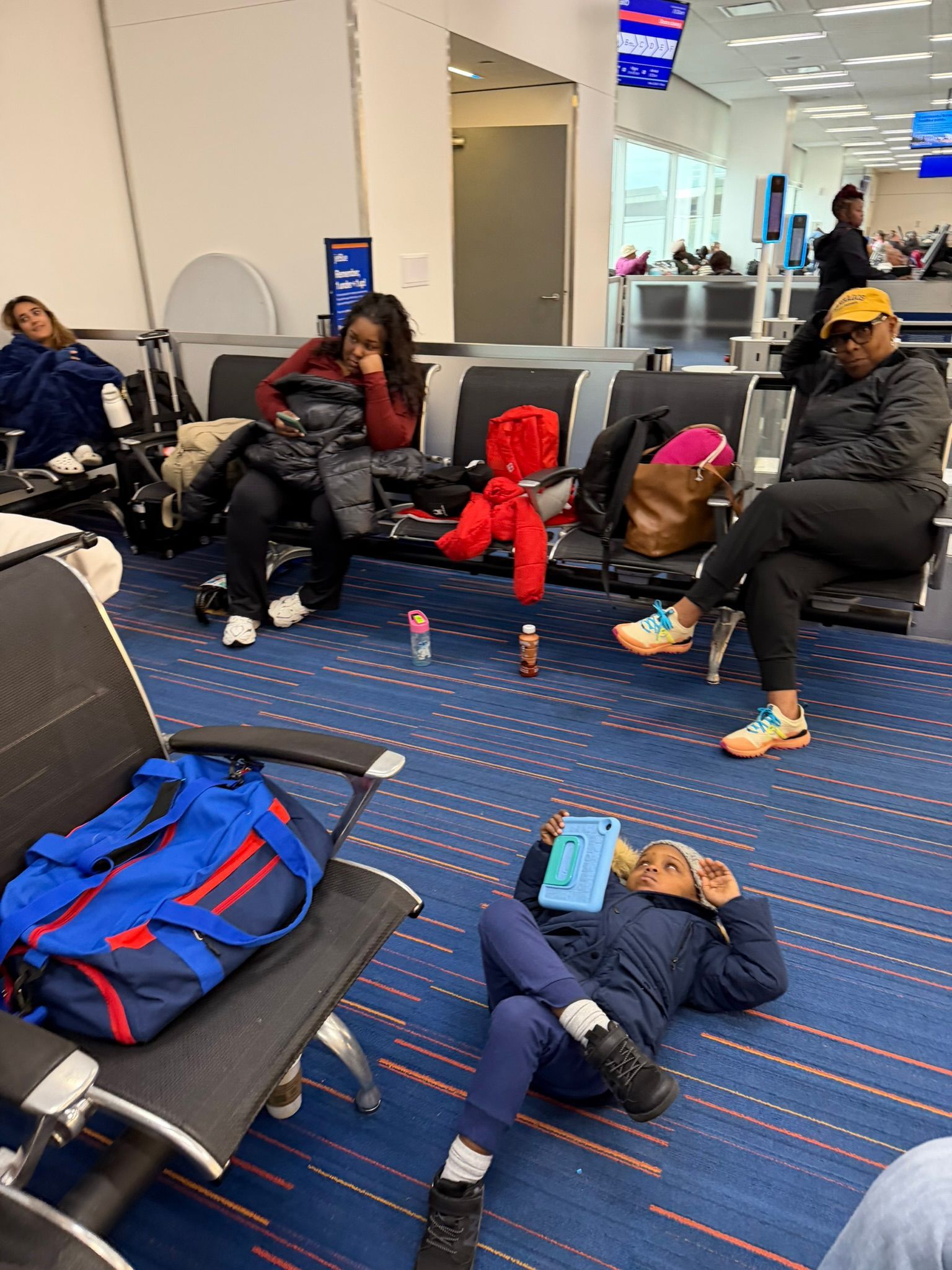 People resting on airport lounge chairs and floor near a gate, with bags and a bright red backpack.
