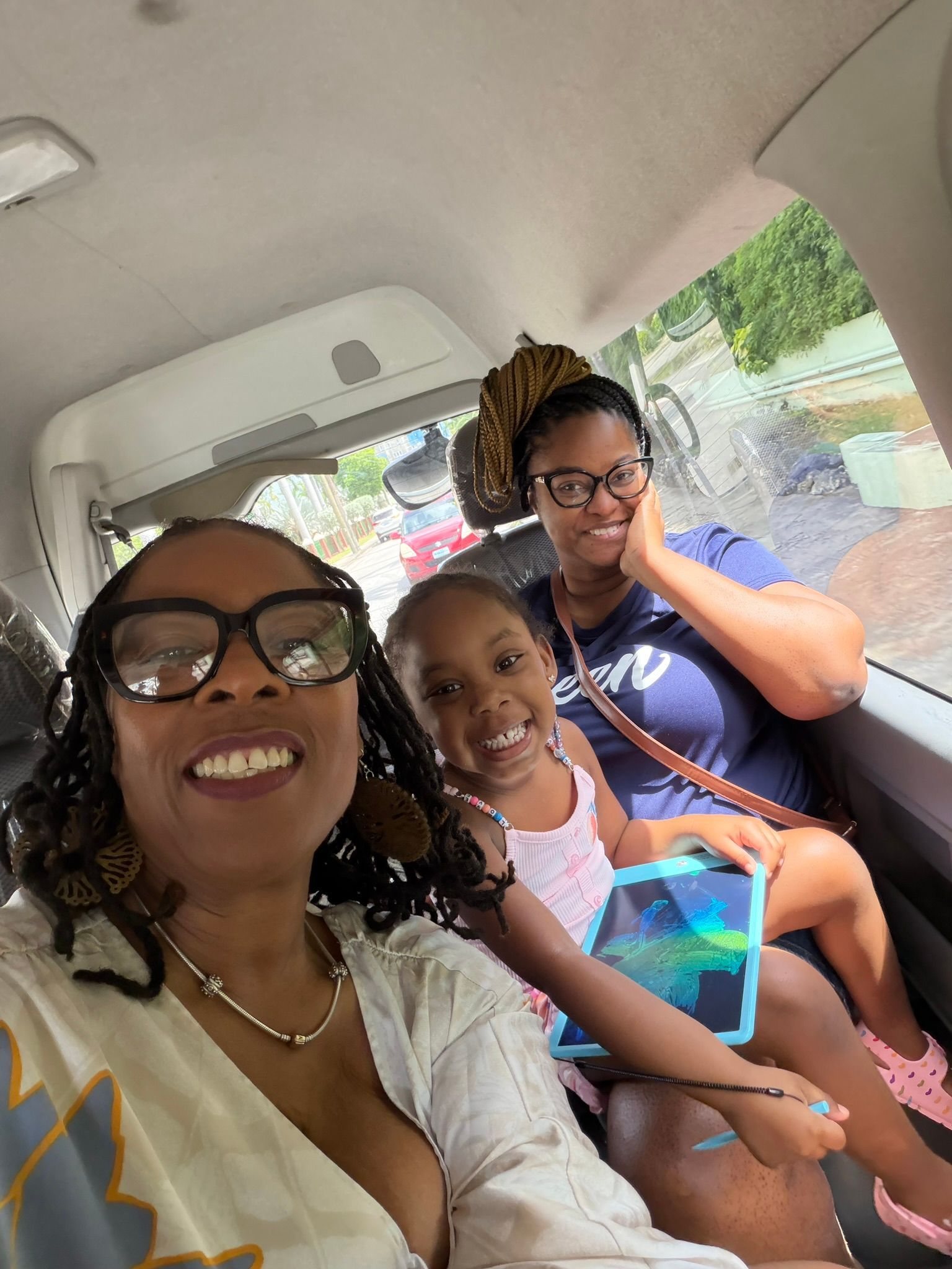 Three people smiling in the back seat of a car, with one holding a tablet.