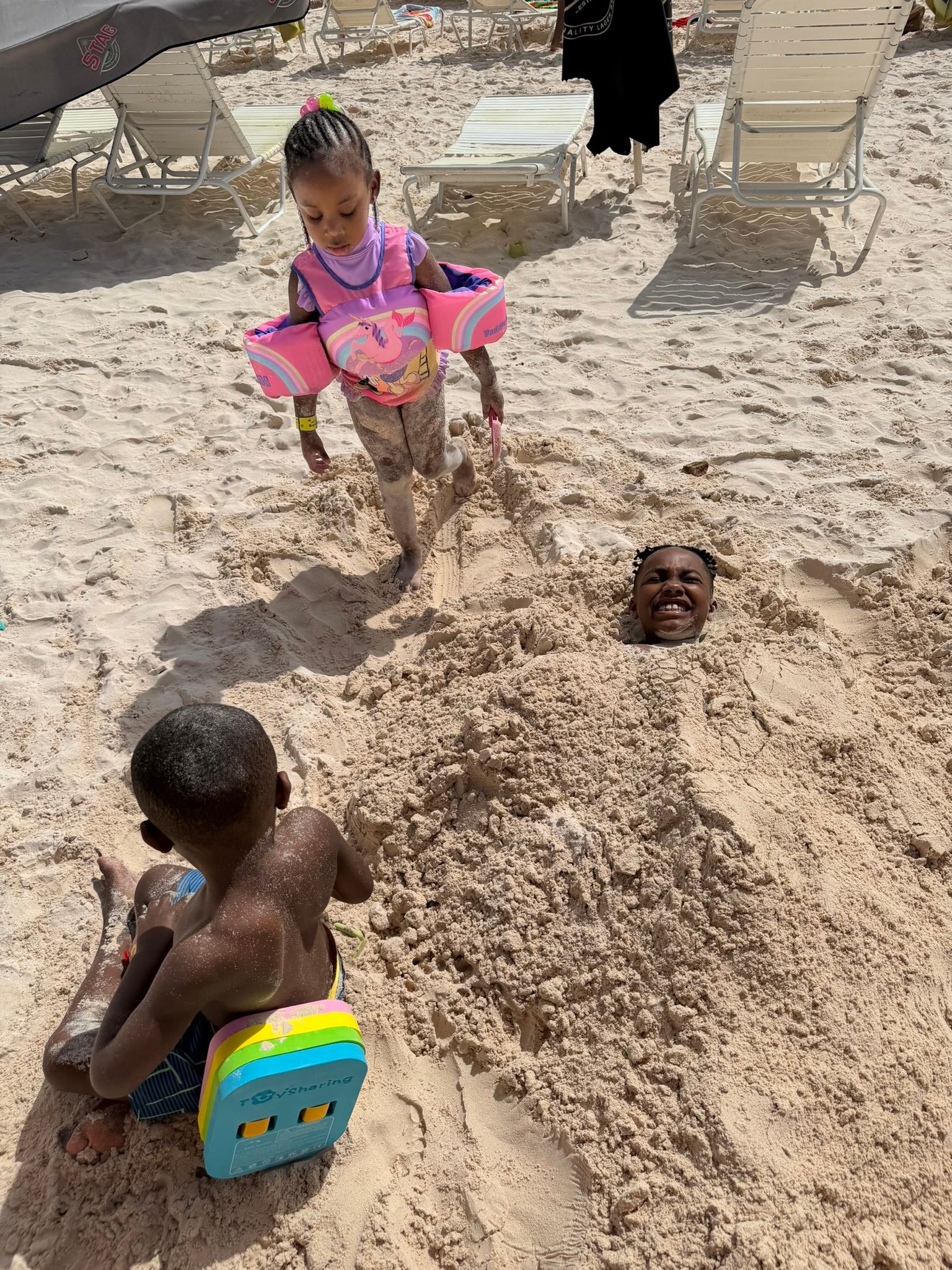 Children playing in sand at a beach, one sitting on a colorful toy seat and another in a pink floatation vest