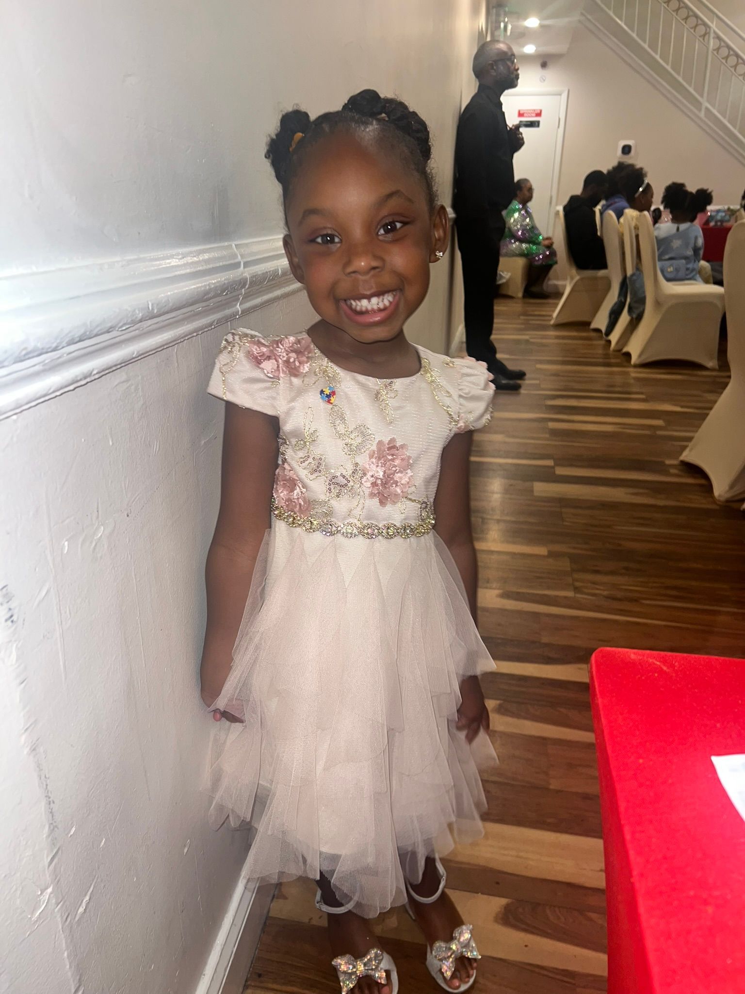 Smiling child in a pink dress standing beside a white wall at an indoor event
