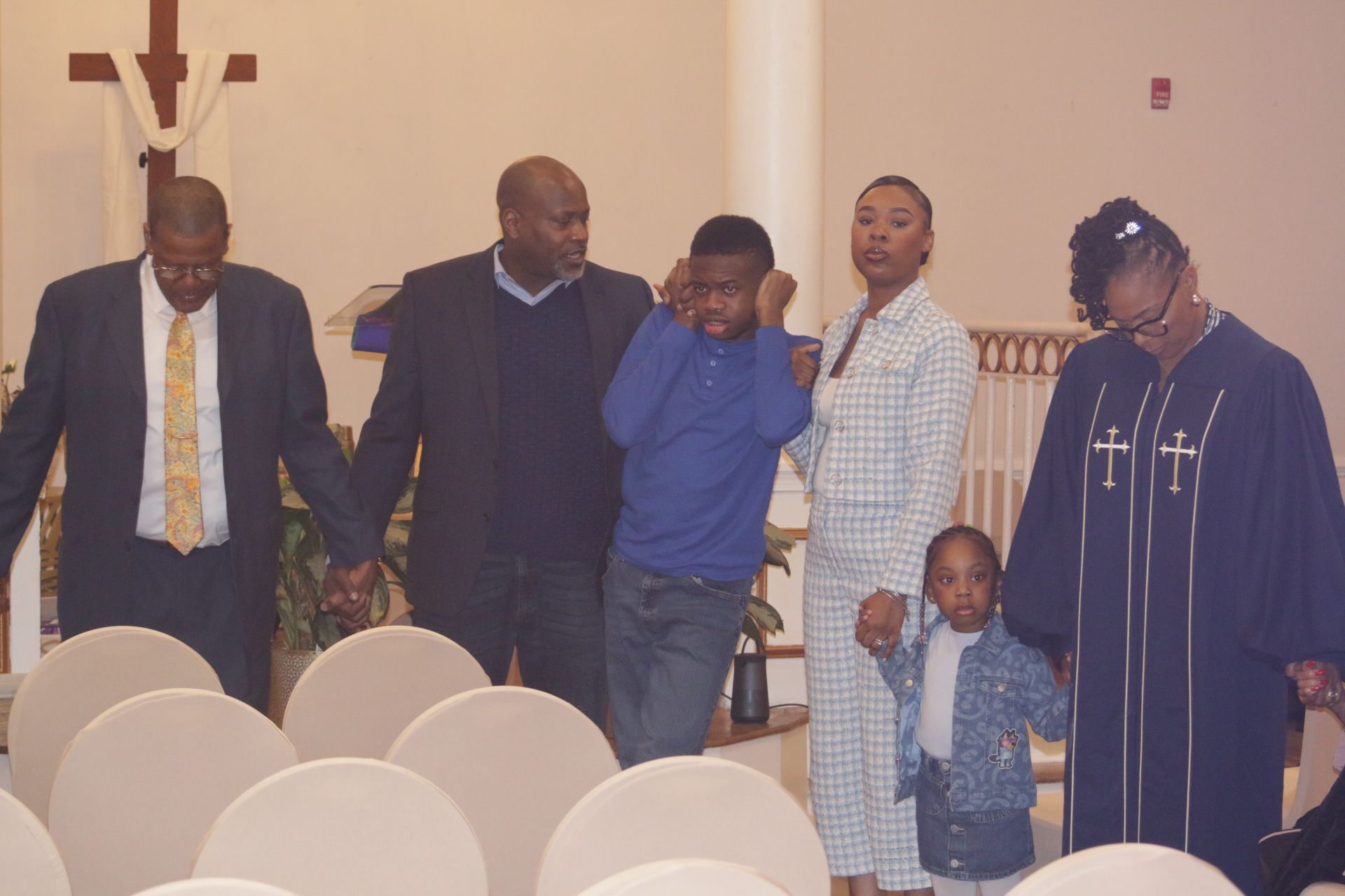 Several people standing in a church aisle, with chairs in the foreground and one child in blue near the center.