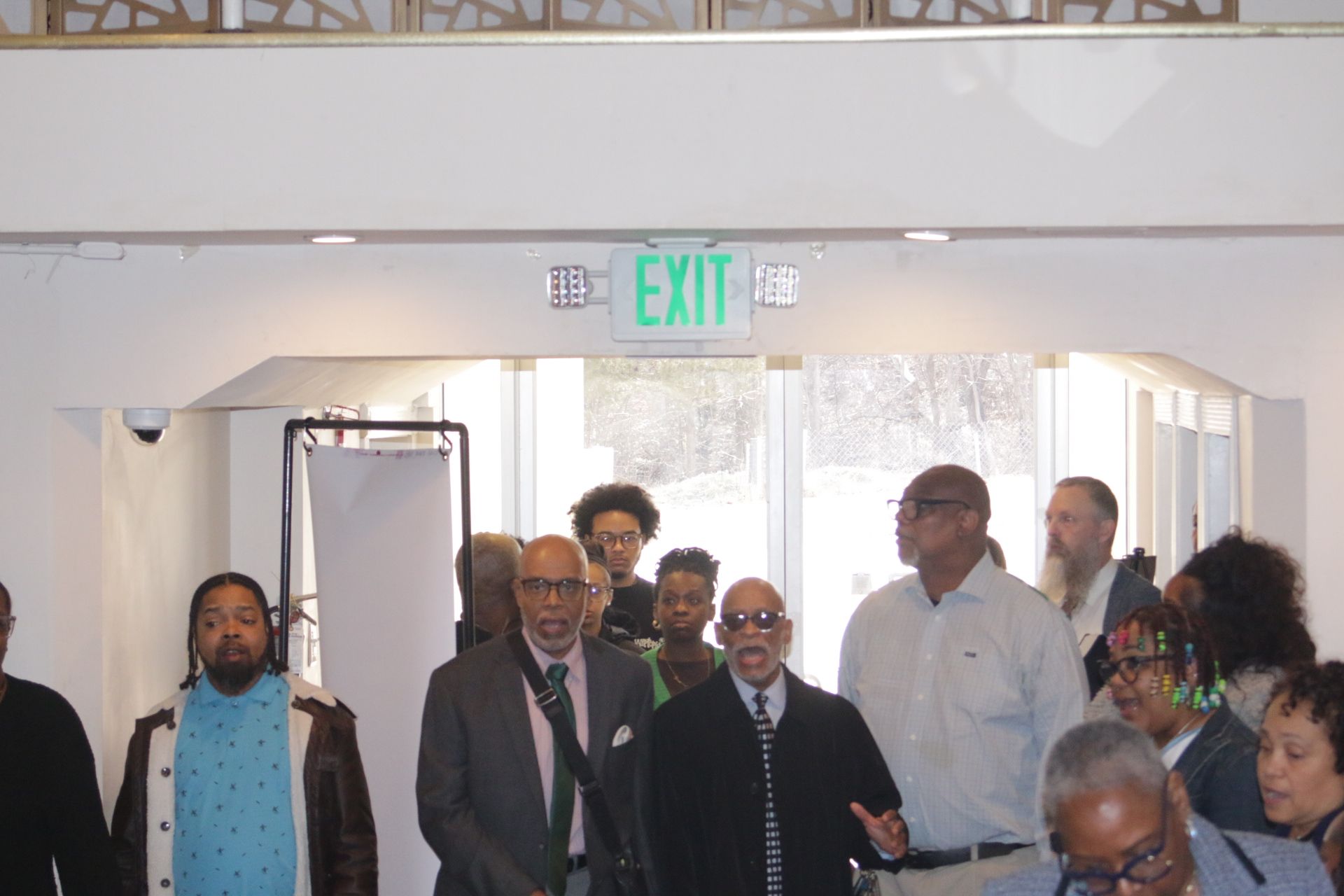People gathered under an EXIT sign in a bright indoor hallway, walking toward the camera.