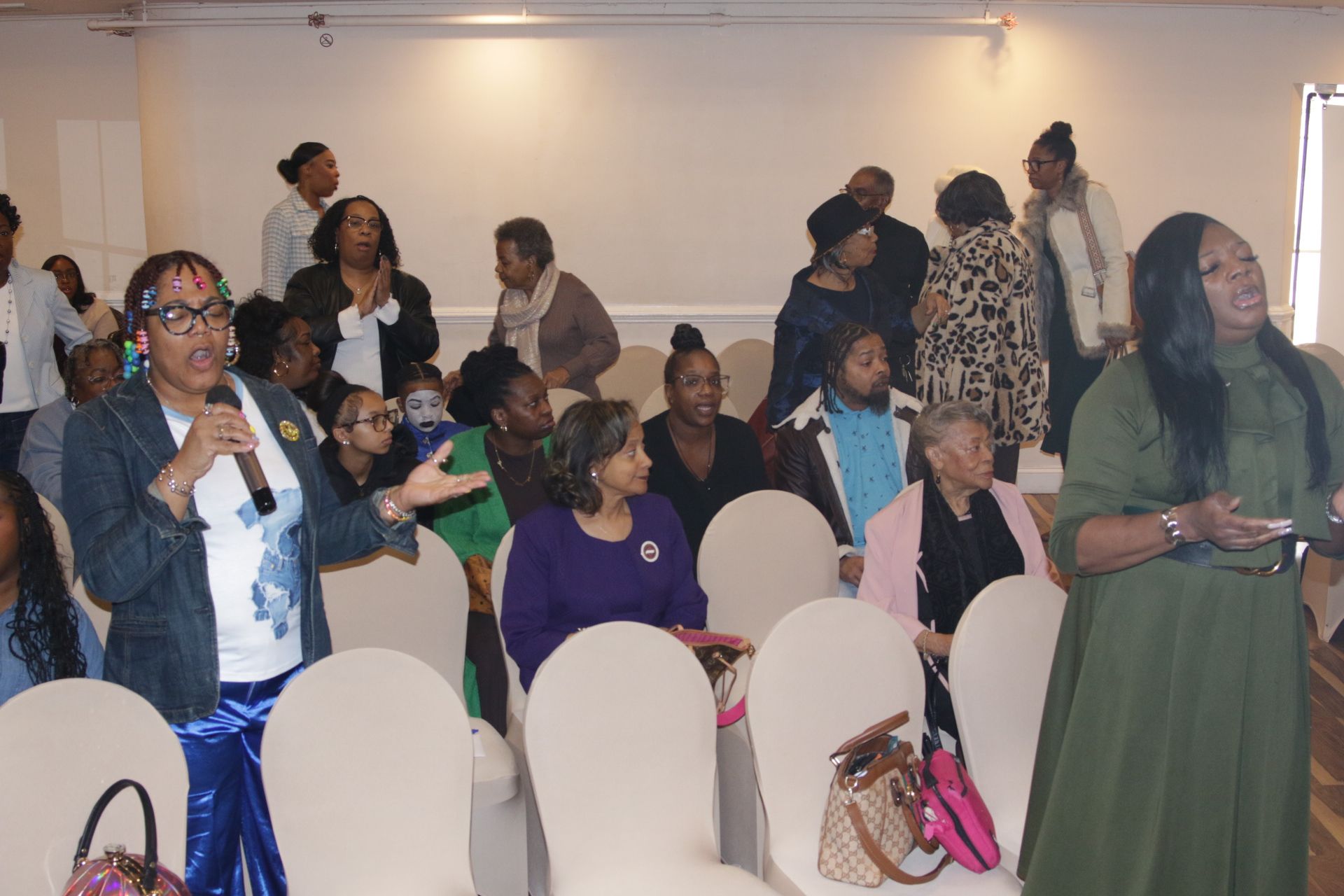 Audience seated in a conference room, with several people standing and talking near the back.
