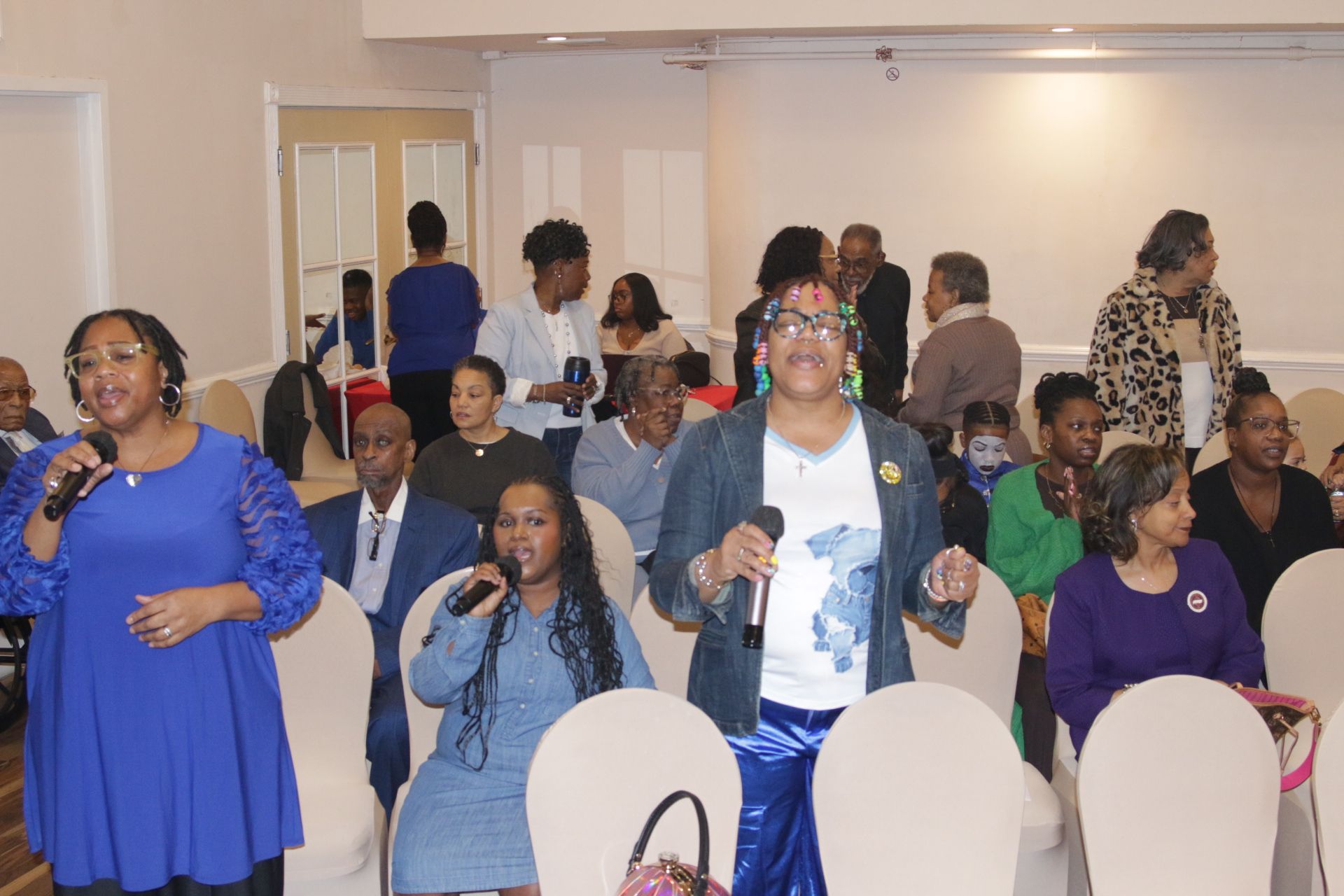 People standing and sitting in a banquet hall, some clapping, with white-covered chairs and warm lighting.