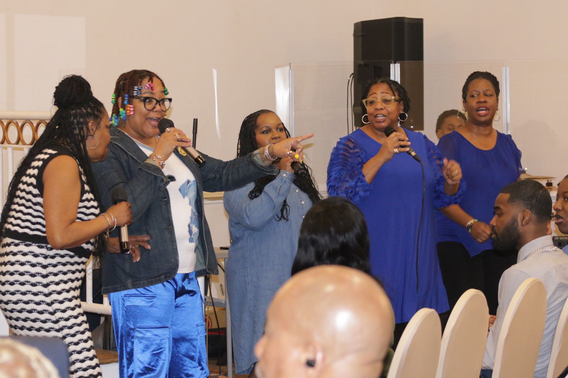 Group of people singing into microphones at an indoor event, with audience seating in the foreground.