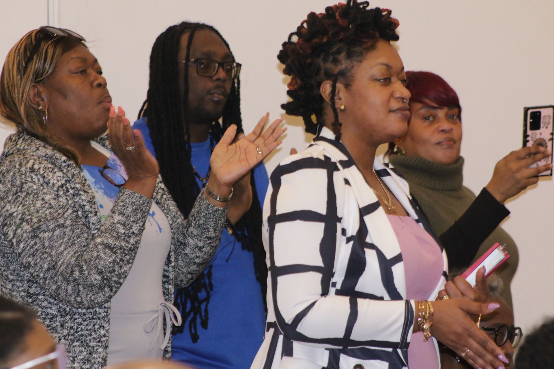 Four people stand indoors, clapping and singing during a gathering; one holds a phone up to record.