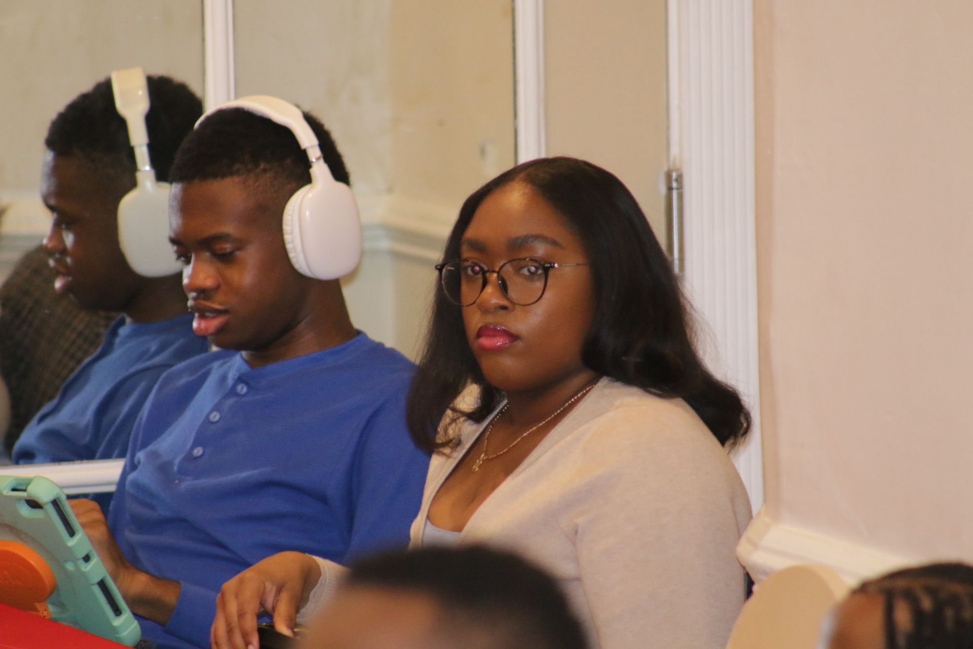 Two people seated indoors, one wearing white headphones and looking at a laptop, the other looking toward the camera.