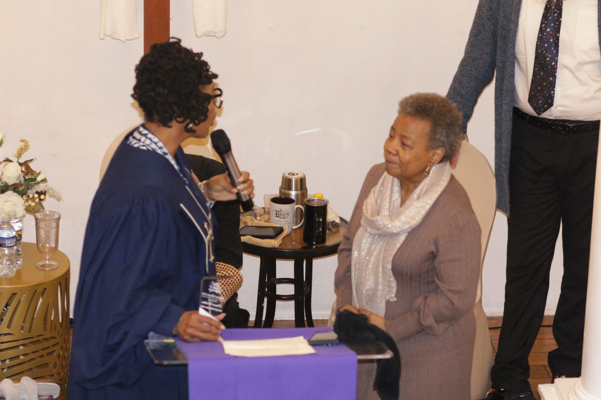 Two people speaking at a small indoor gathering beside a table with drinks and flowers.
