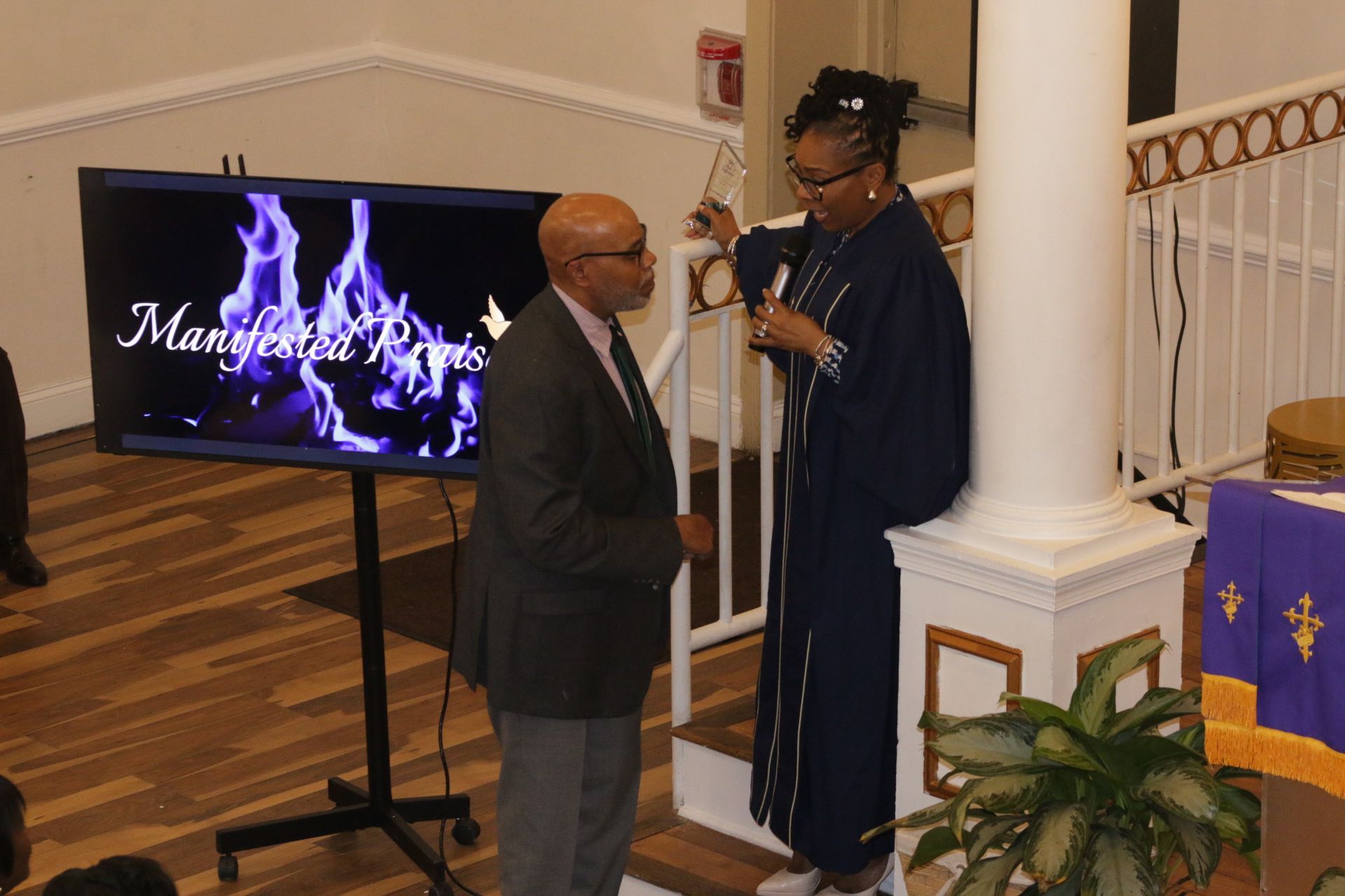 Two people speak at a church podium beside a lit screen showing flames.