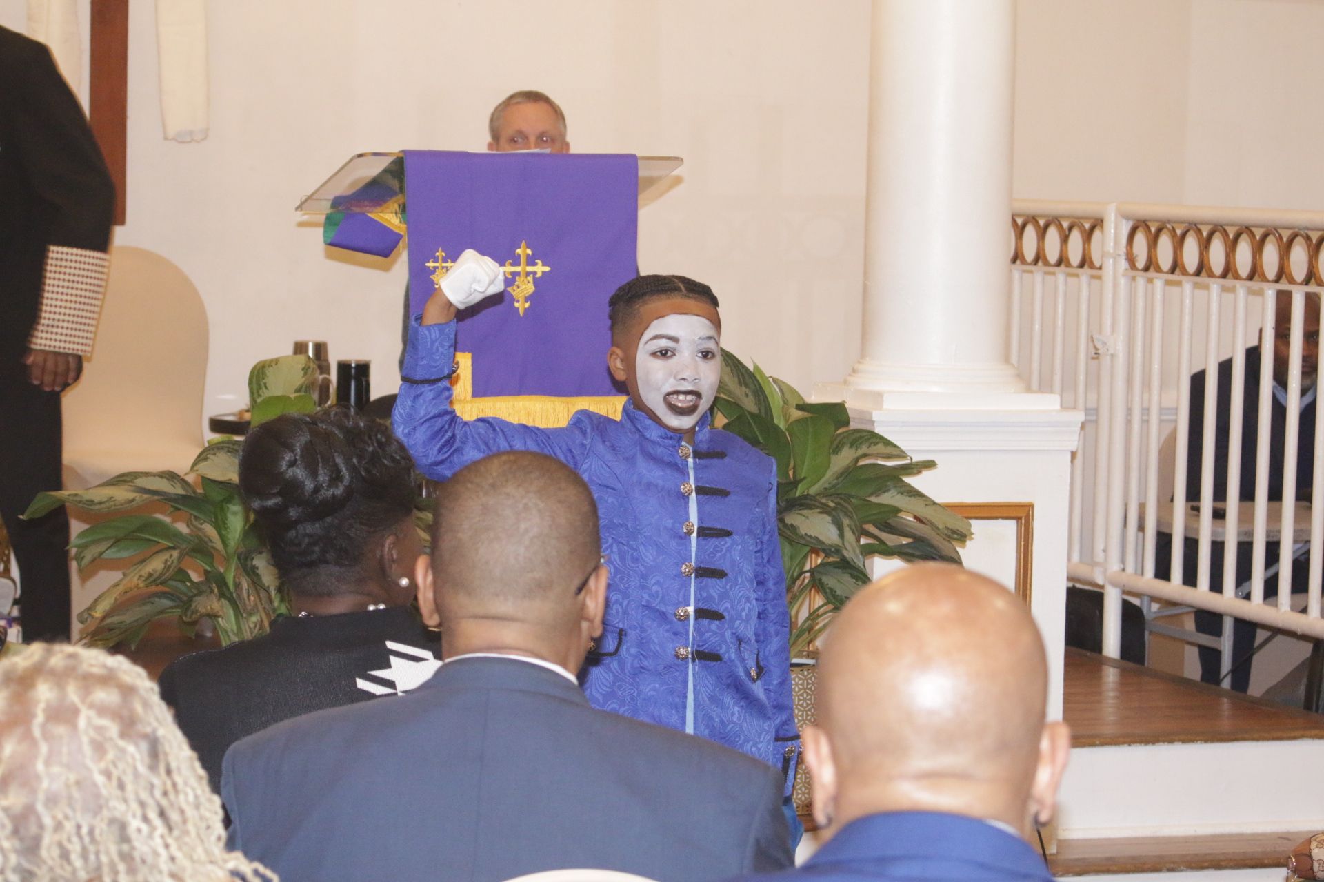 Person in blue robe speaking at a podium in a church, with audience seated in front.