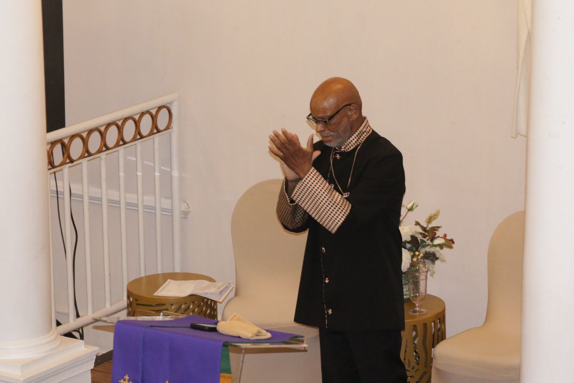 Man in black jacket clapping beside a purple-covered table in a church-like room