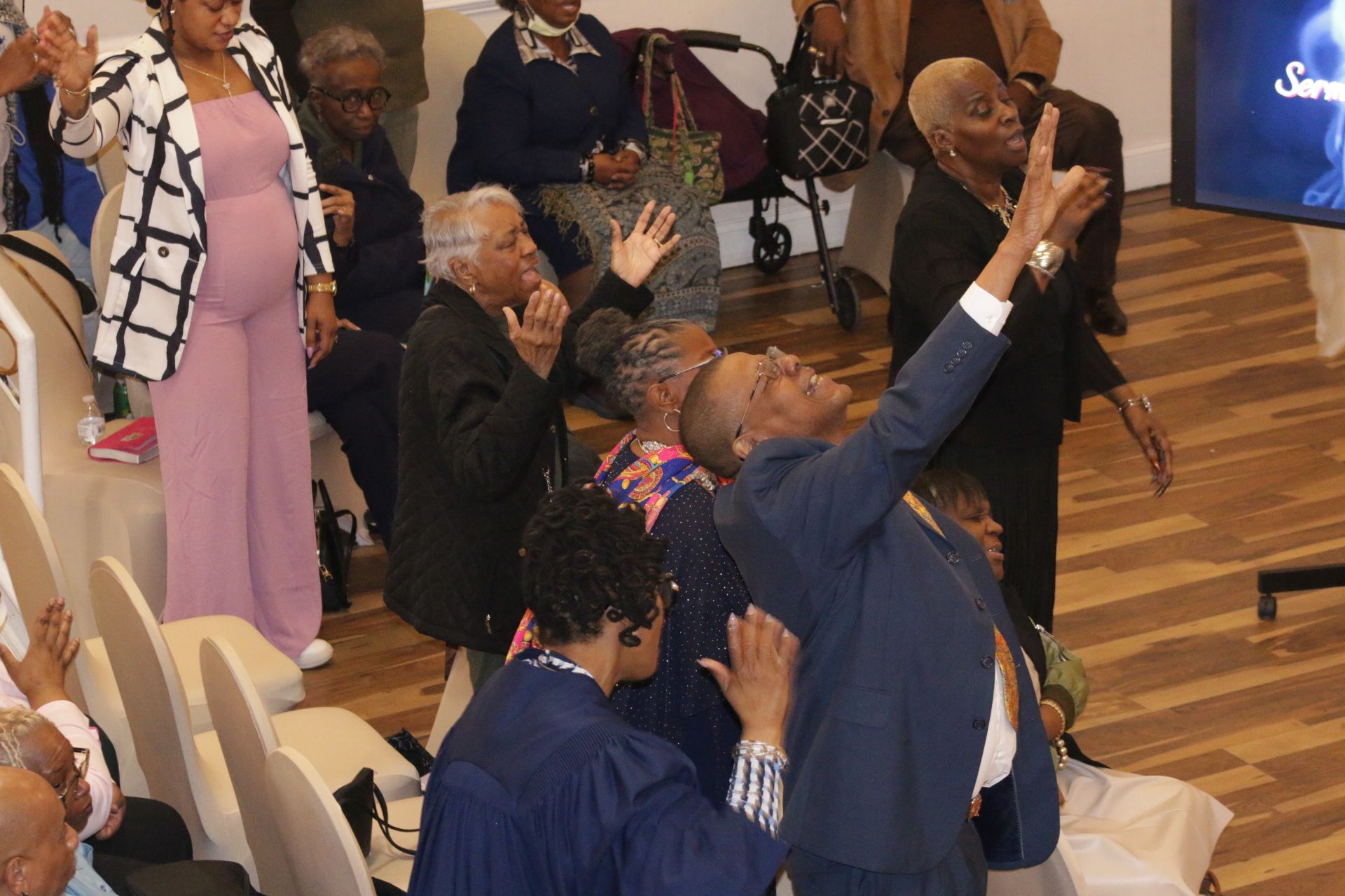 People in church raising hands in worship, with seated attendees and a wooden floor.