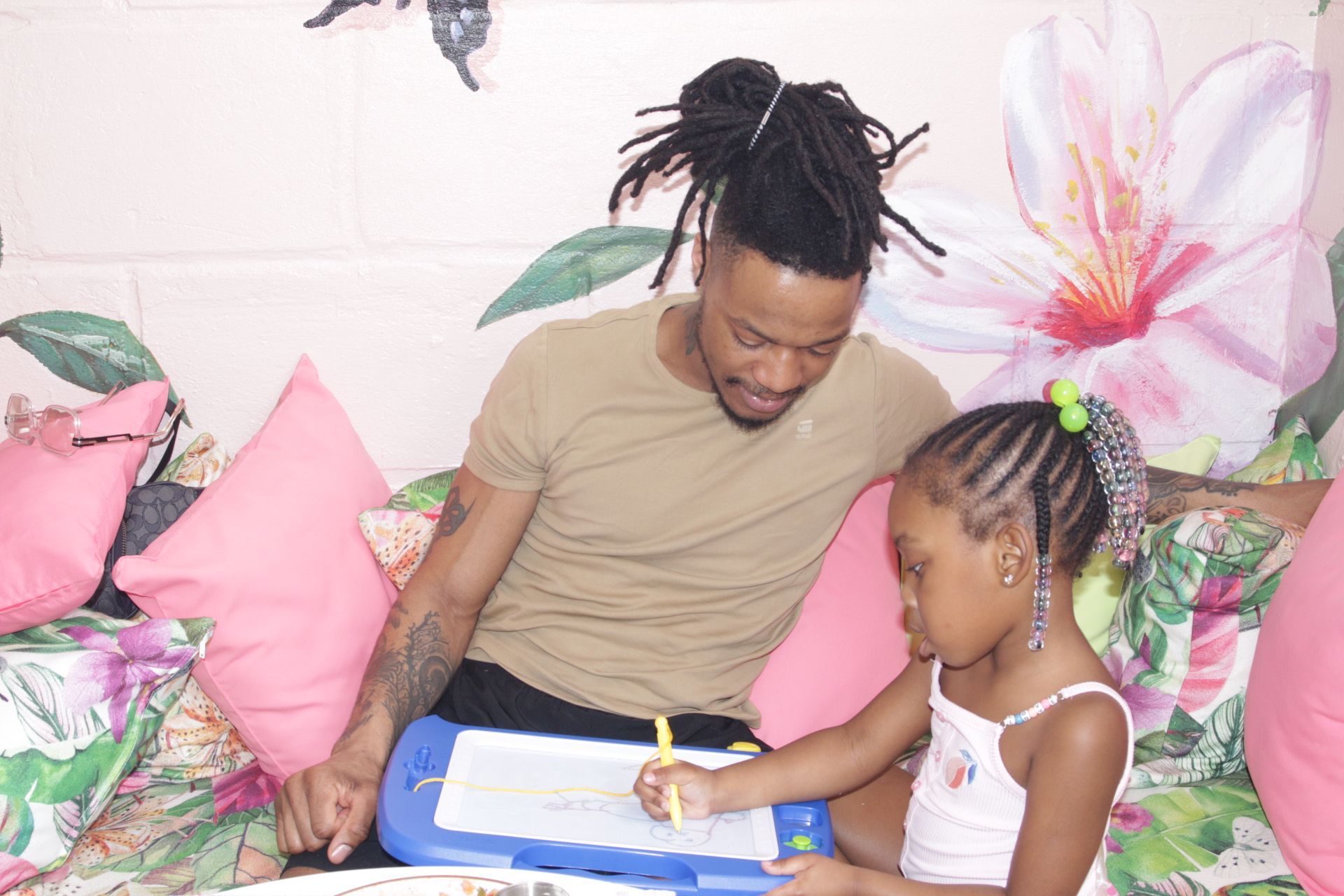 Man and child writing together on a blue clipboard in a colorful room with pink pillows and flowers