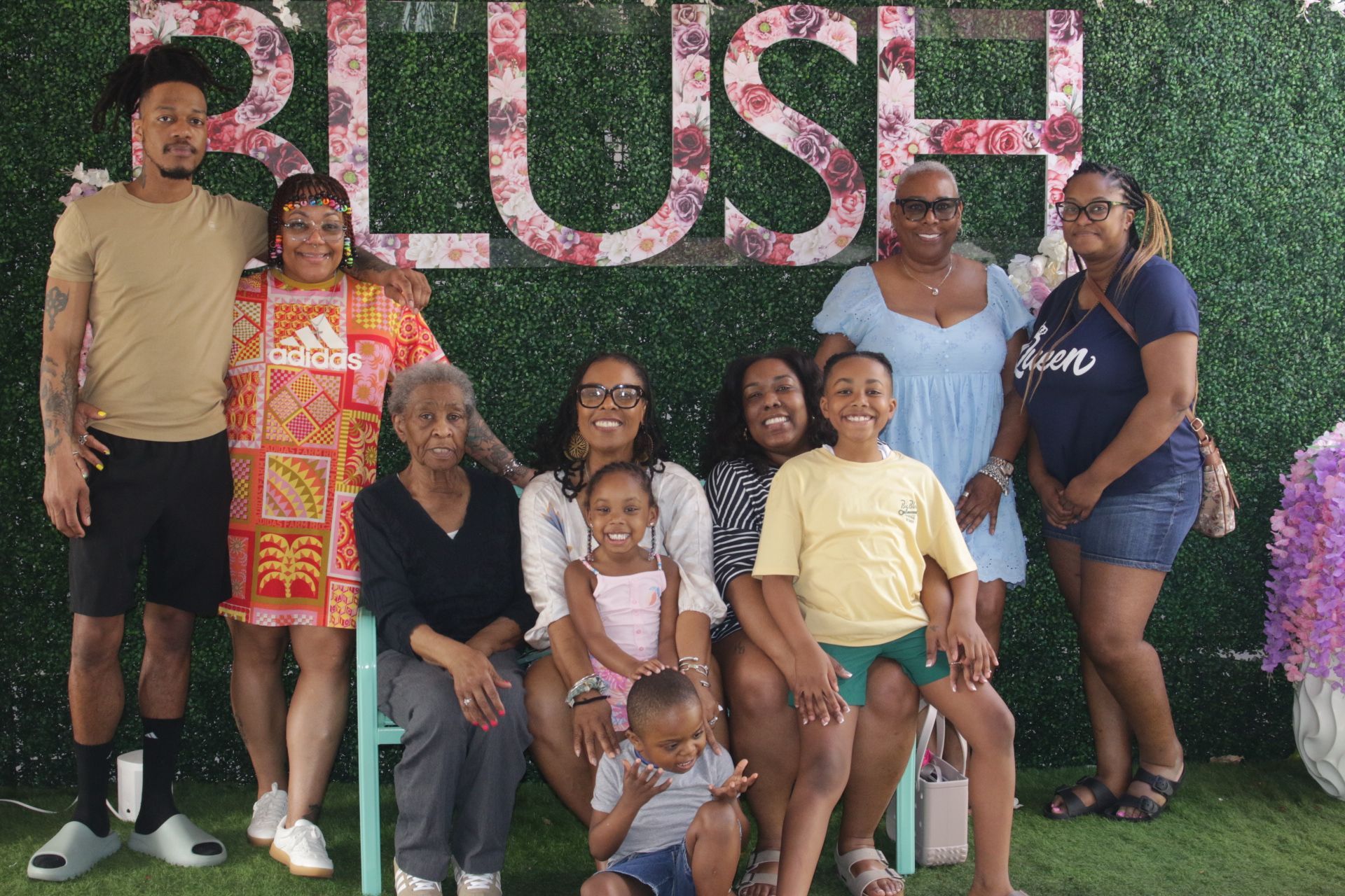 Group of seven posing in front of a “RUSH” floral wall, smiling outdoors in casual summer clothes.