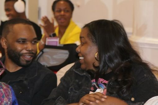 Two people smiling and talking at an indoor event, with a woman in a yellow jacket in the background