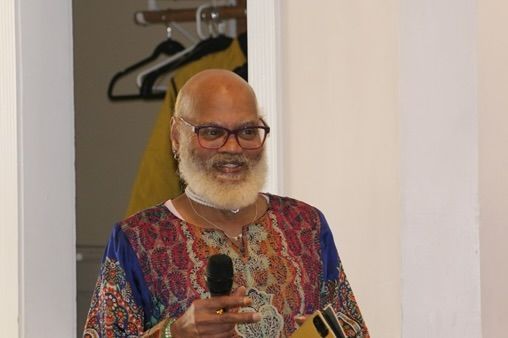 Man smiling and holding a microphone indoors, wearing a colorful patterned shirt.