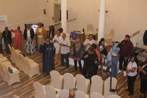 People standing in line inside a church or hall with rows of beige chairs in the foreground