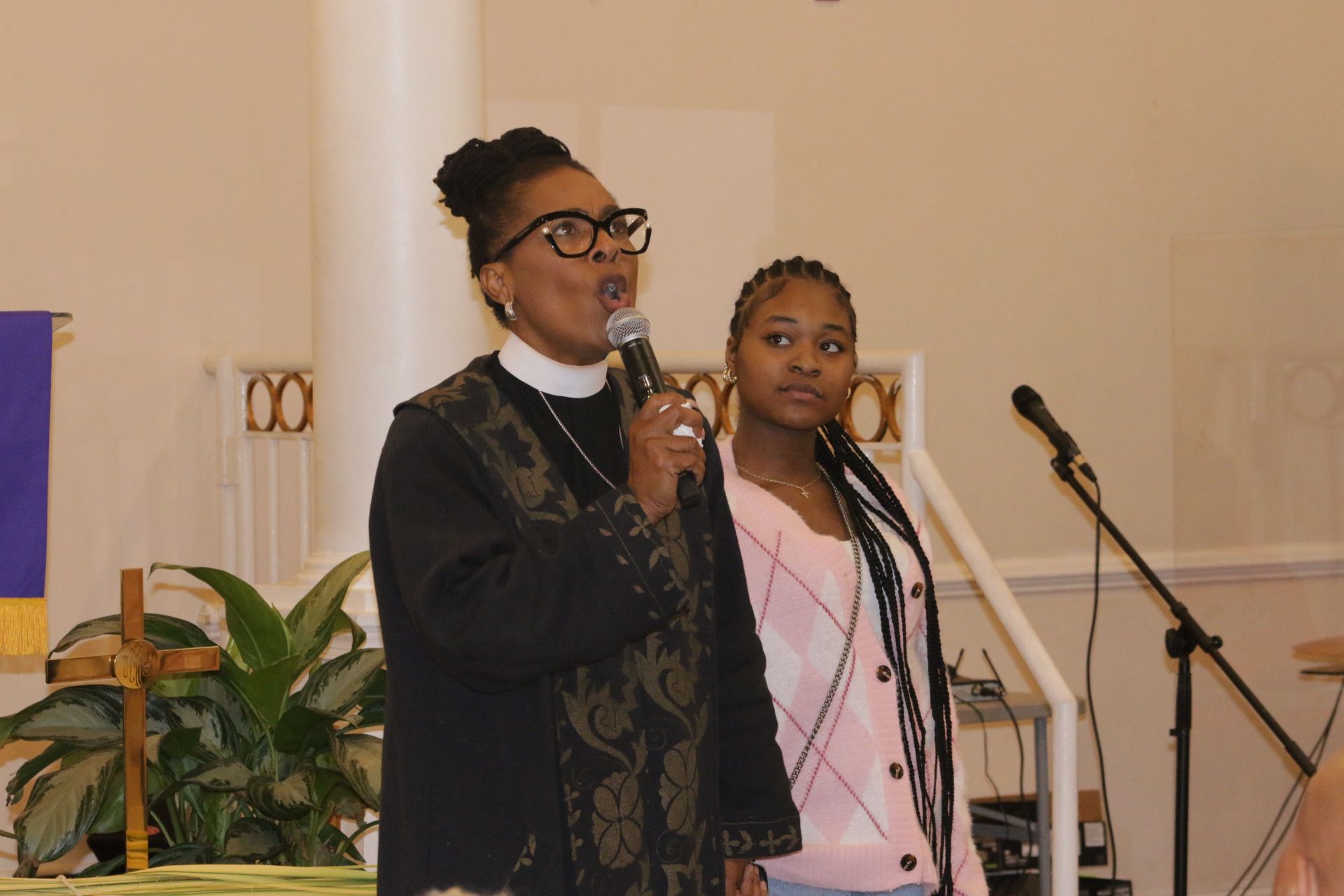 Two people speaking at microphones in a church or hall, with potted plants and stairs behind them.