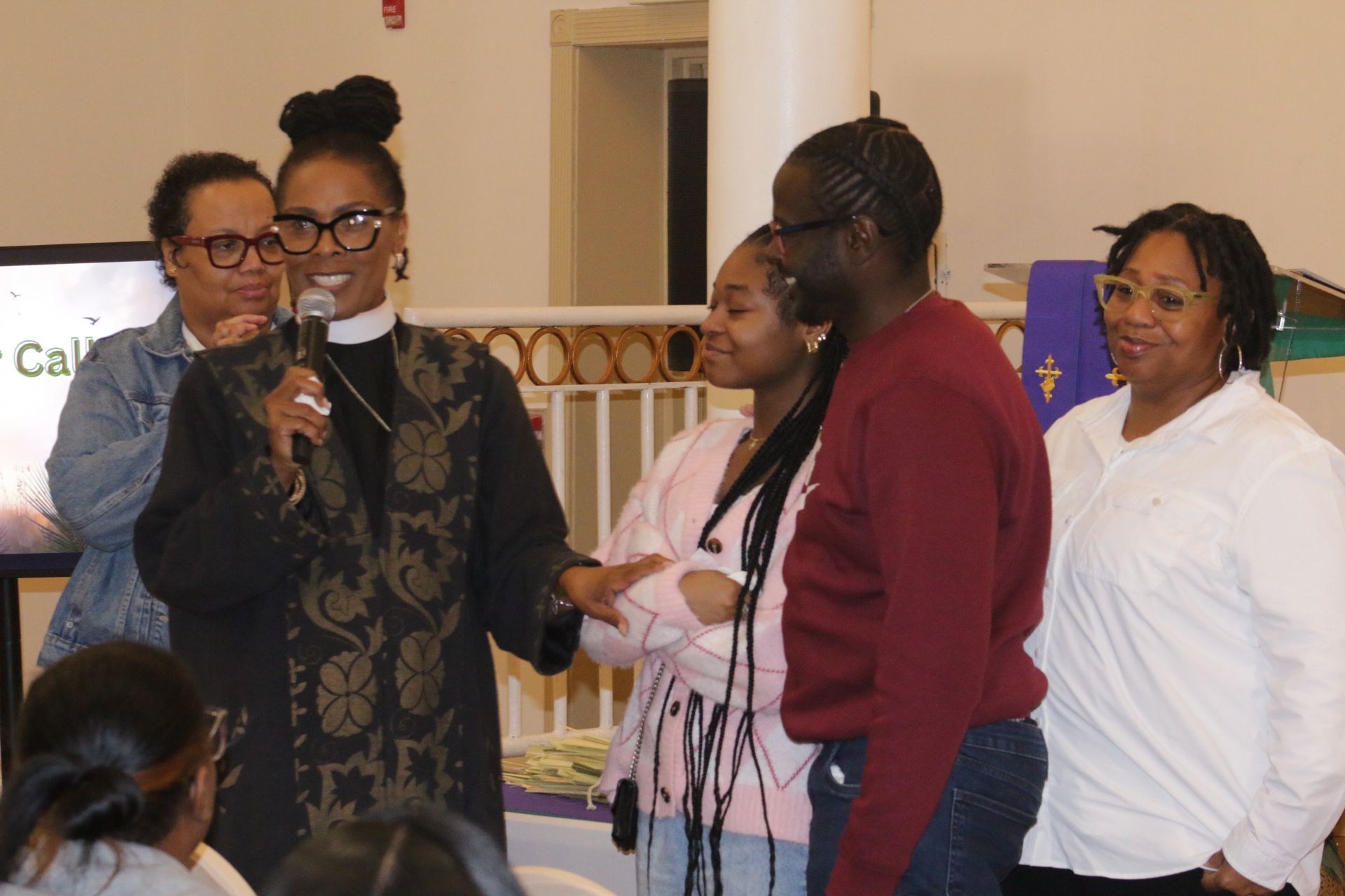 People at an indoor event, one speaking into a microphone while others stand nearby and listen.