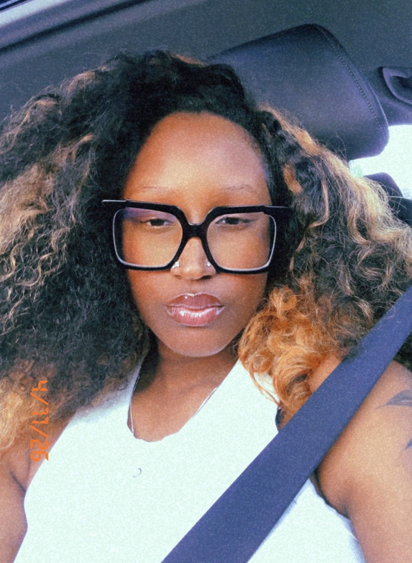 Person wearing black glasses and a seatbelt in a car, with curly hair and a white top.