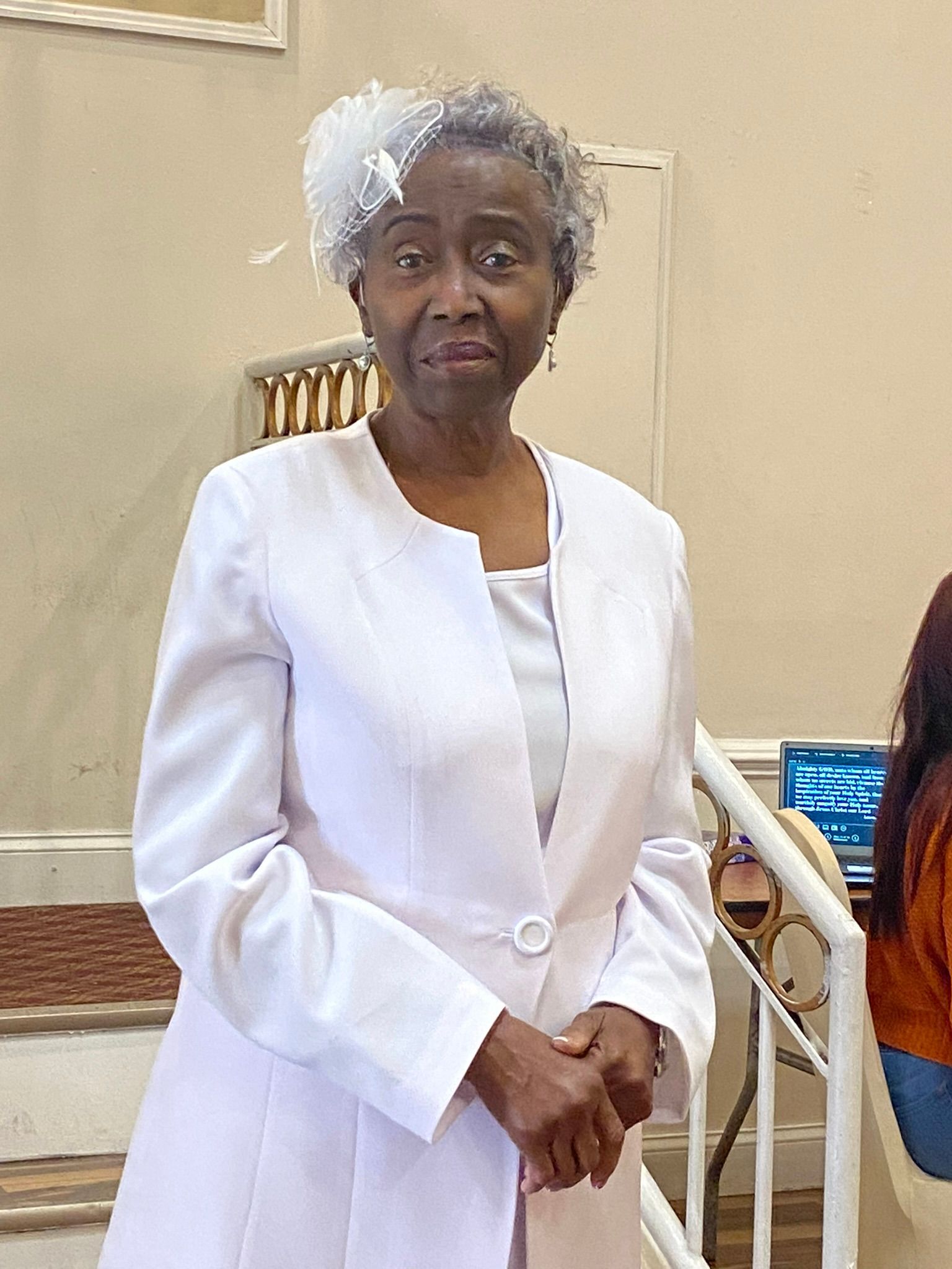 Woman in a white outfit standing indoors with hands clasped, near a wall and staircase.