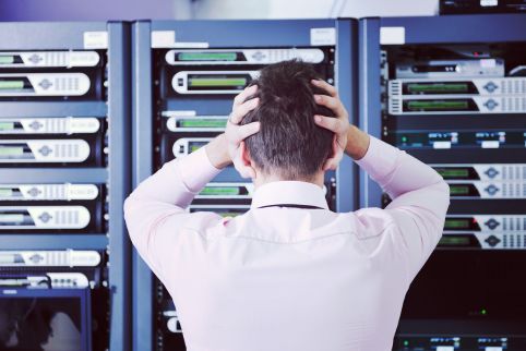 A man in a pink shirt is holding his head in front of a server rack.