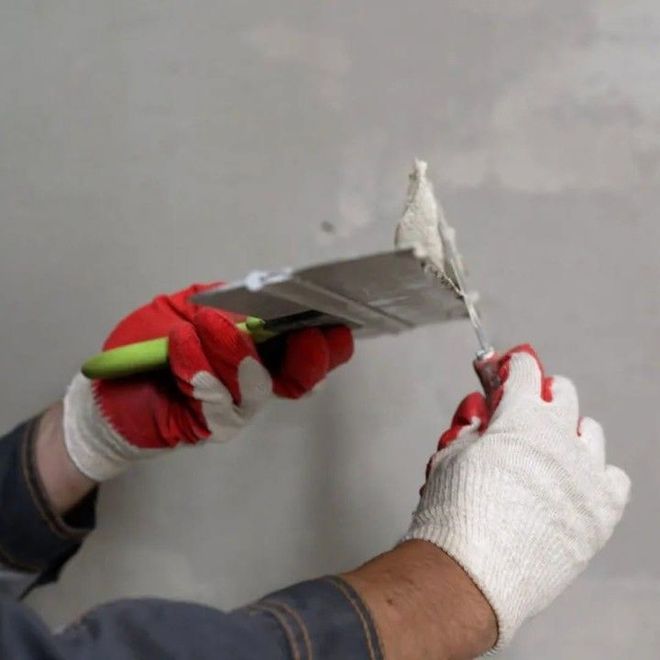 Gloved hands using a putty knife to scrape and apply plaster on a wall