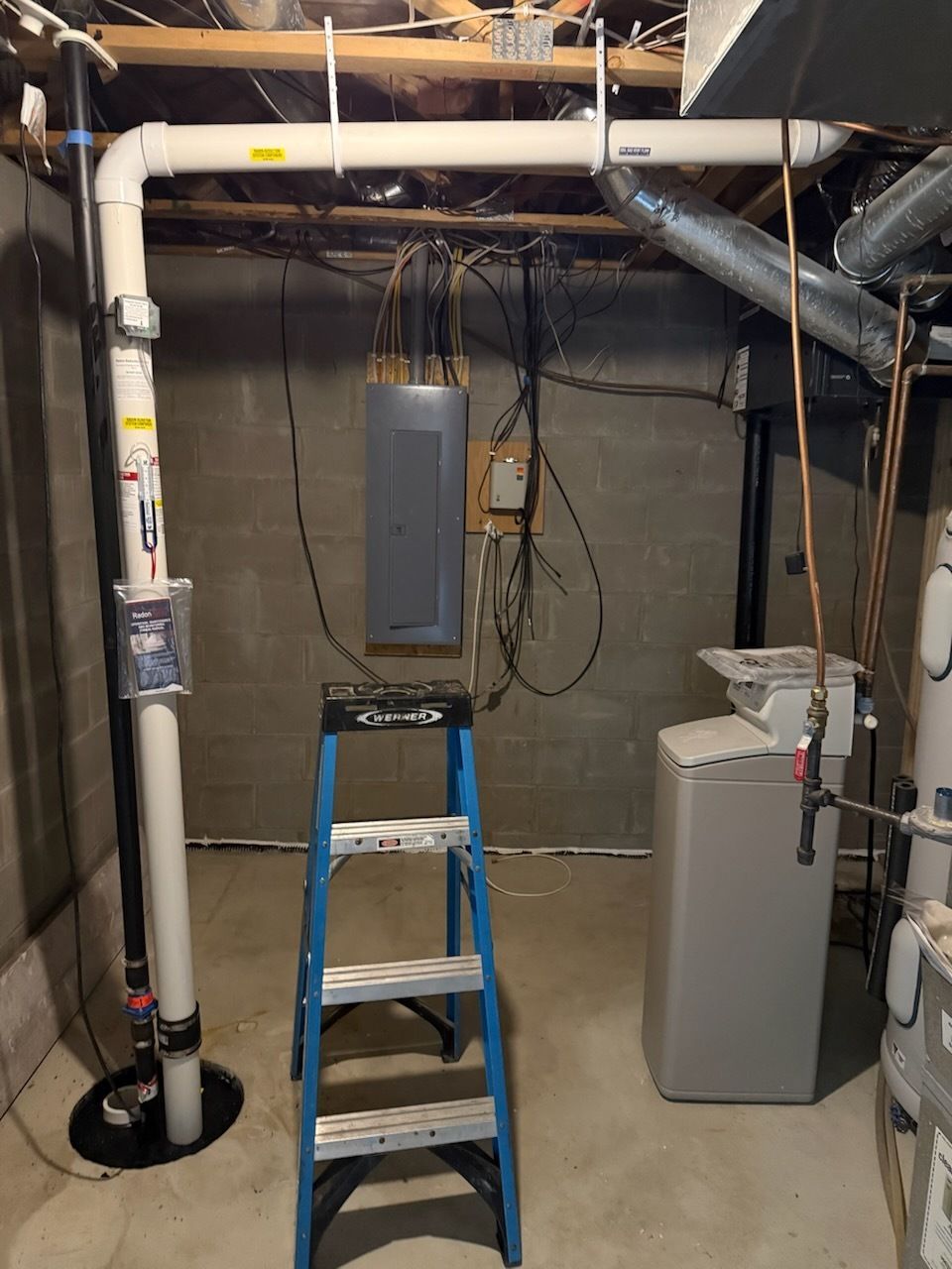 Basement utility area with a blue step ladder under exposed pipes and electrical panels.