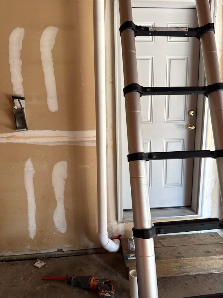 Ladder and PVC pipes in a home repair area beside a patched wall with exposed drywall and tools on the floor