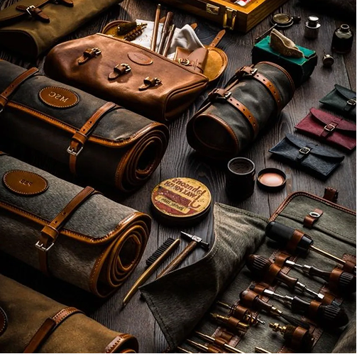 Assorted leather travel gear and grooming tools arranged on a dark table, in brown, black, and brass tones