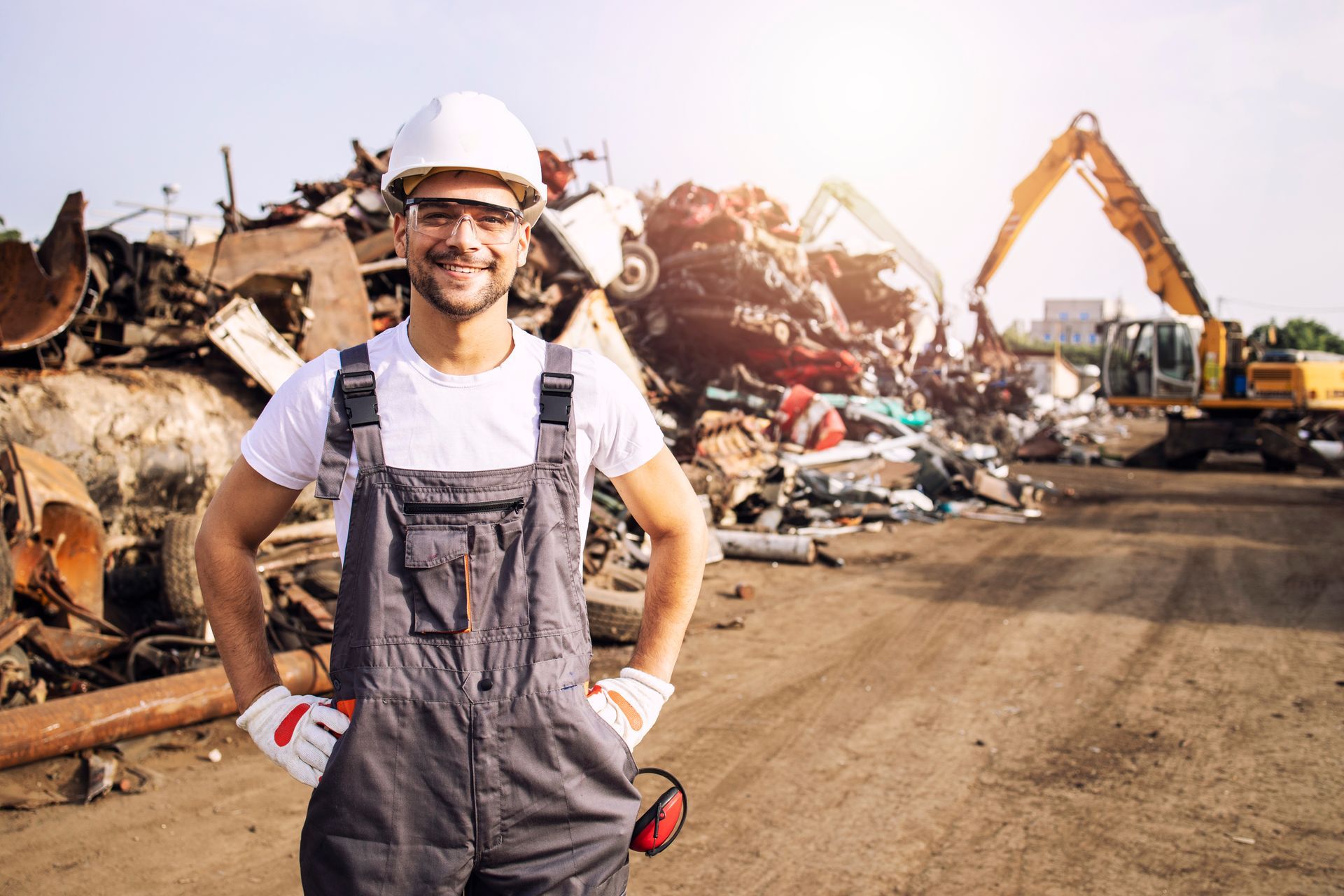 Worker in hard hat and overalls at a scrapyard with metal piles and an excavator. Worker in hard hat and overalls at a scrapyard with metal piles and an excavator.