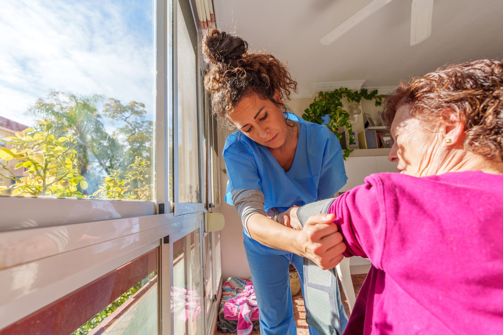 Caregiver assisting a person with movement; blue scrubs, pink top, sunny balcony setting.