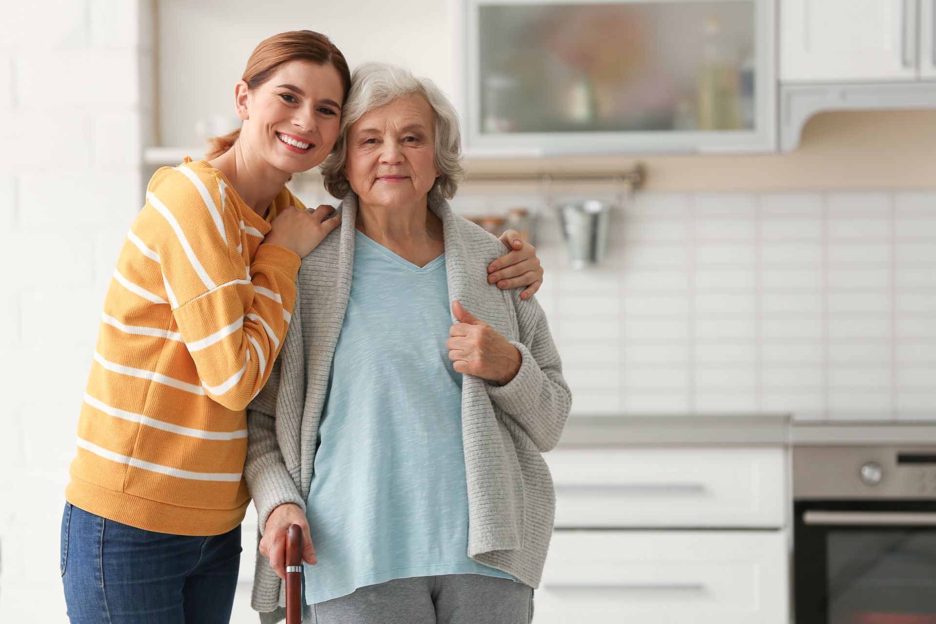 Woman in yellow sweater hugs older woman with a cane in a kitchen. Both smile.