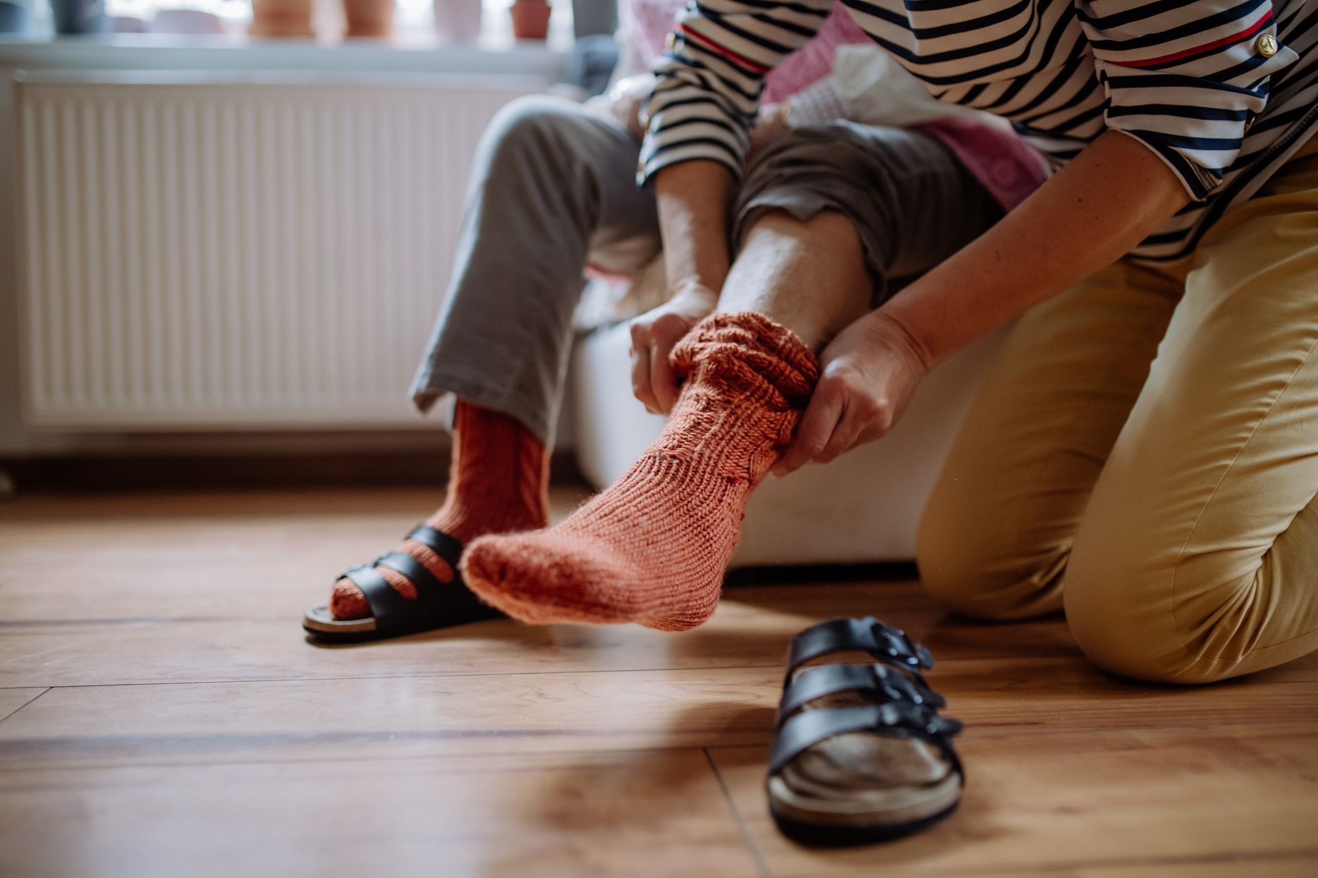 Person helping another put on an orange sock indoors, near a radiator and sandals.