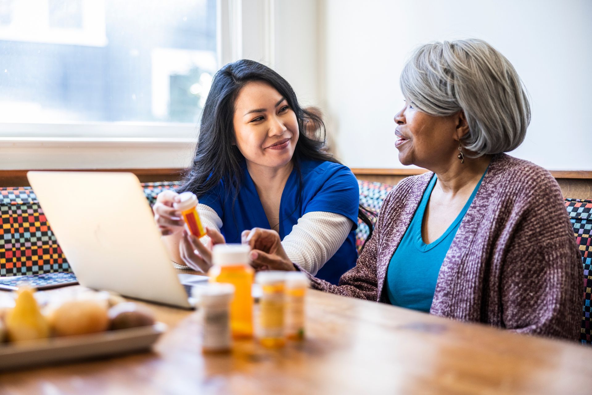 Healthcare worker reviewing medication bottles with a patient, laptop open.