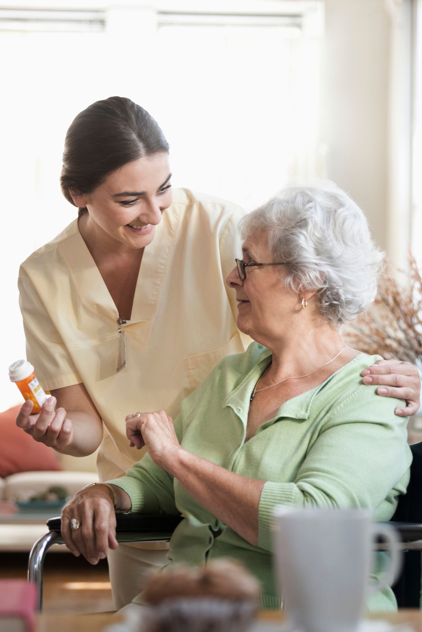 Caregiver handing pills to an older adult in a wheelchair, smiling in a brightly lit room.