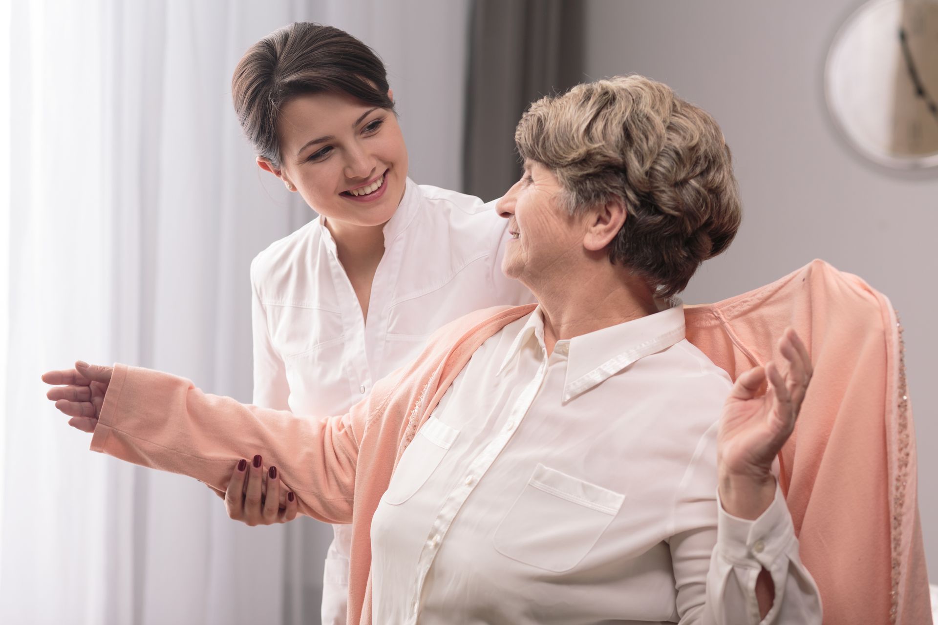 Woman helping another woman put on a pink cardigan in a room with a window; both smiling.