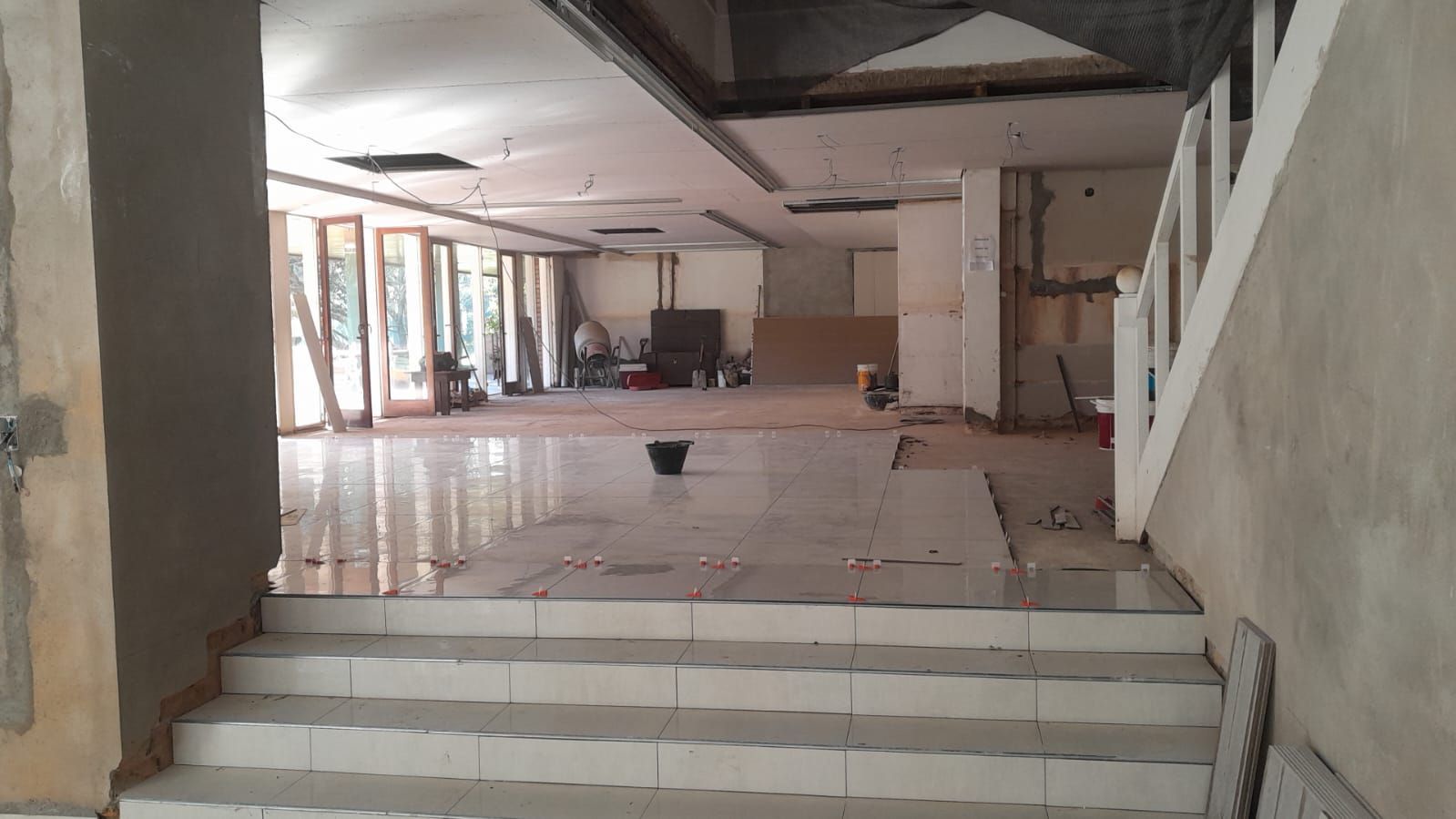 Vestíbulo interior vacío e inacabado, con suelo de baldosas, escalones al frente y vigas del techo a la vista.