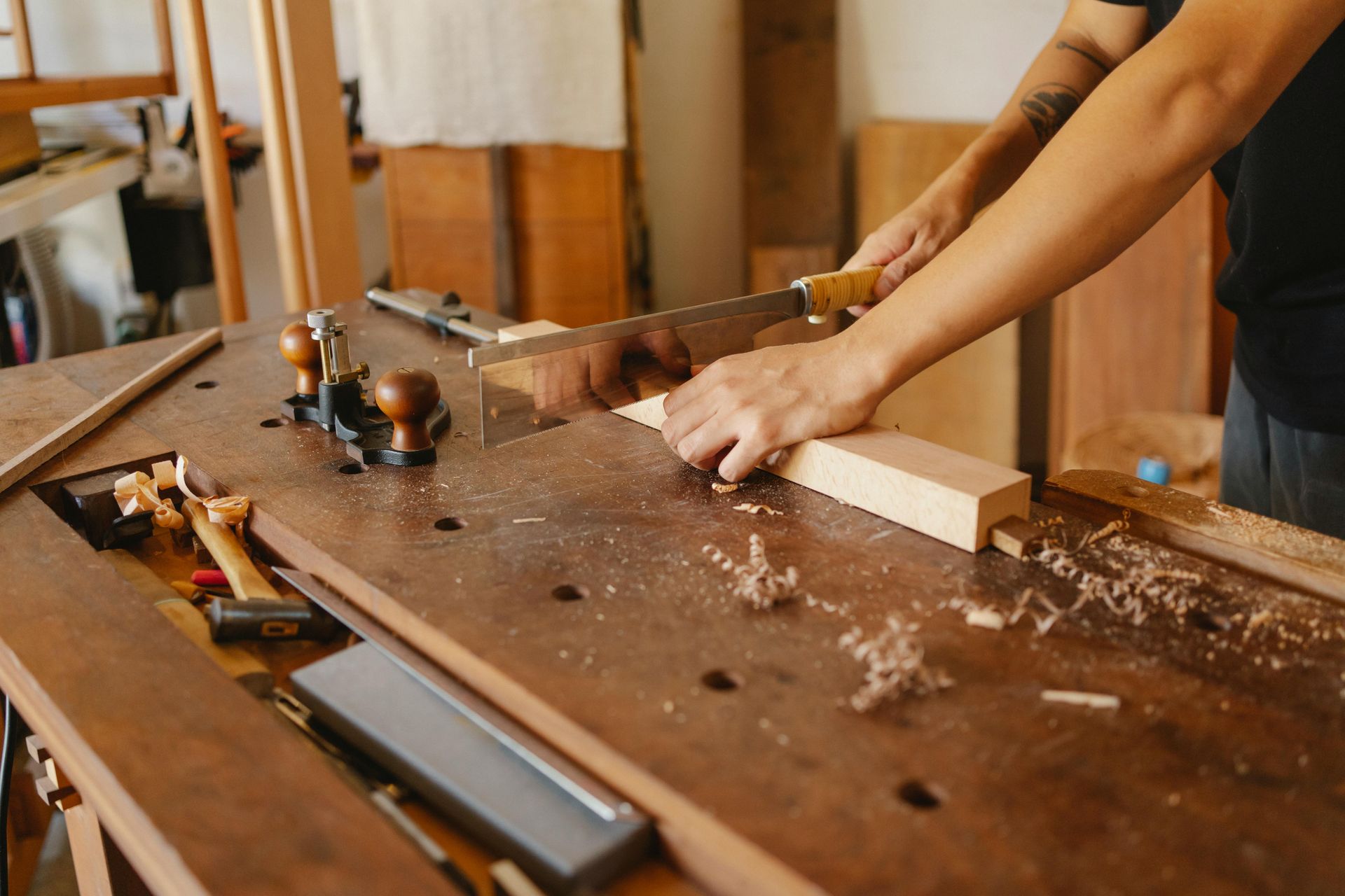 A person uses a handsaw to cut a wooden block on a sturdy workshop workbench surrounded by various tools and wood shavings.