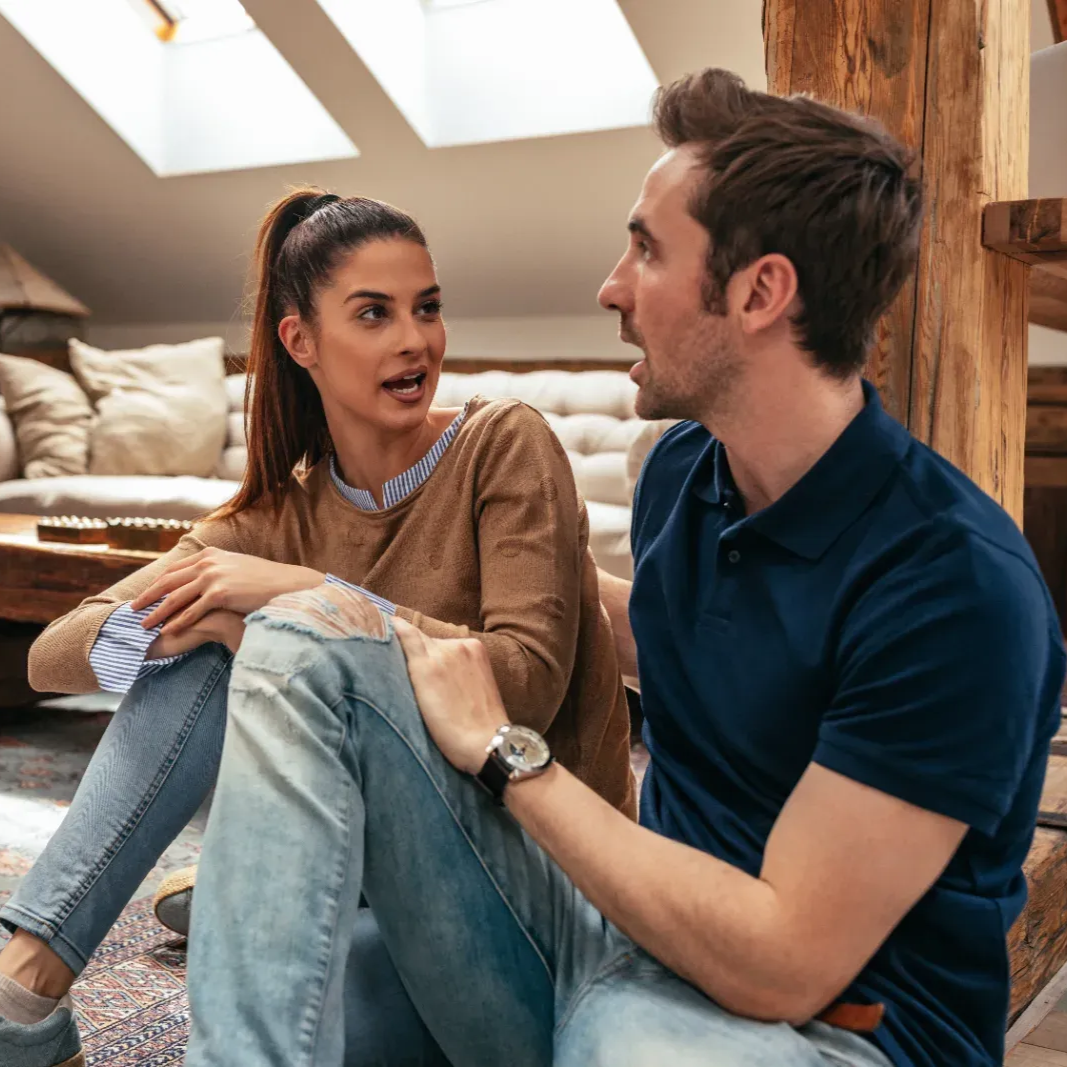 Two people sit on the floor in a bright living room, talking and smiling near a couch.