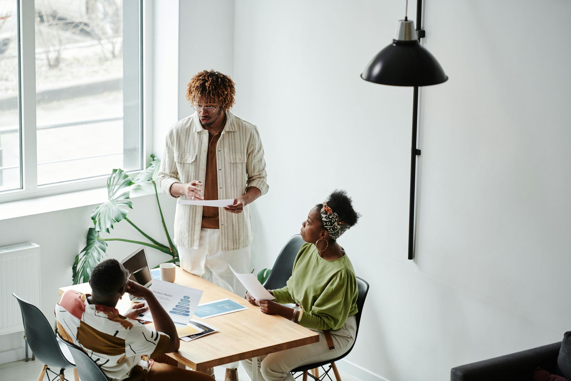 Three people in a bright room reviewing papers. One man stands, two women sit at a table, discussing.