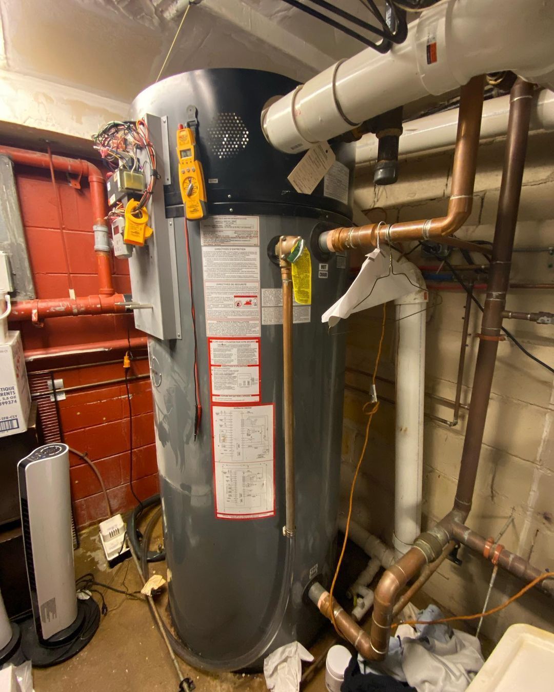 A large water heater is sitting in a basement surrounded by pipes.