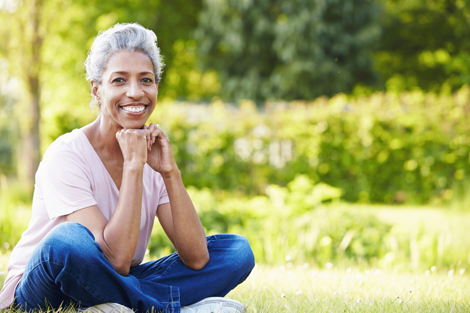 Smiling person sitting cross-legged on grass in a sunny park, with greenery in the background