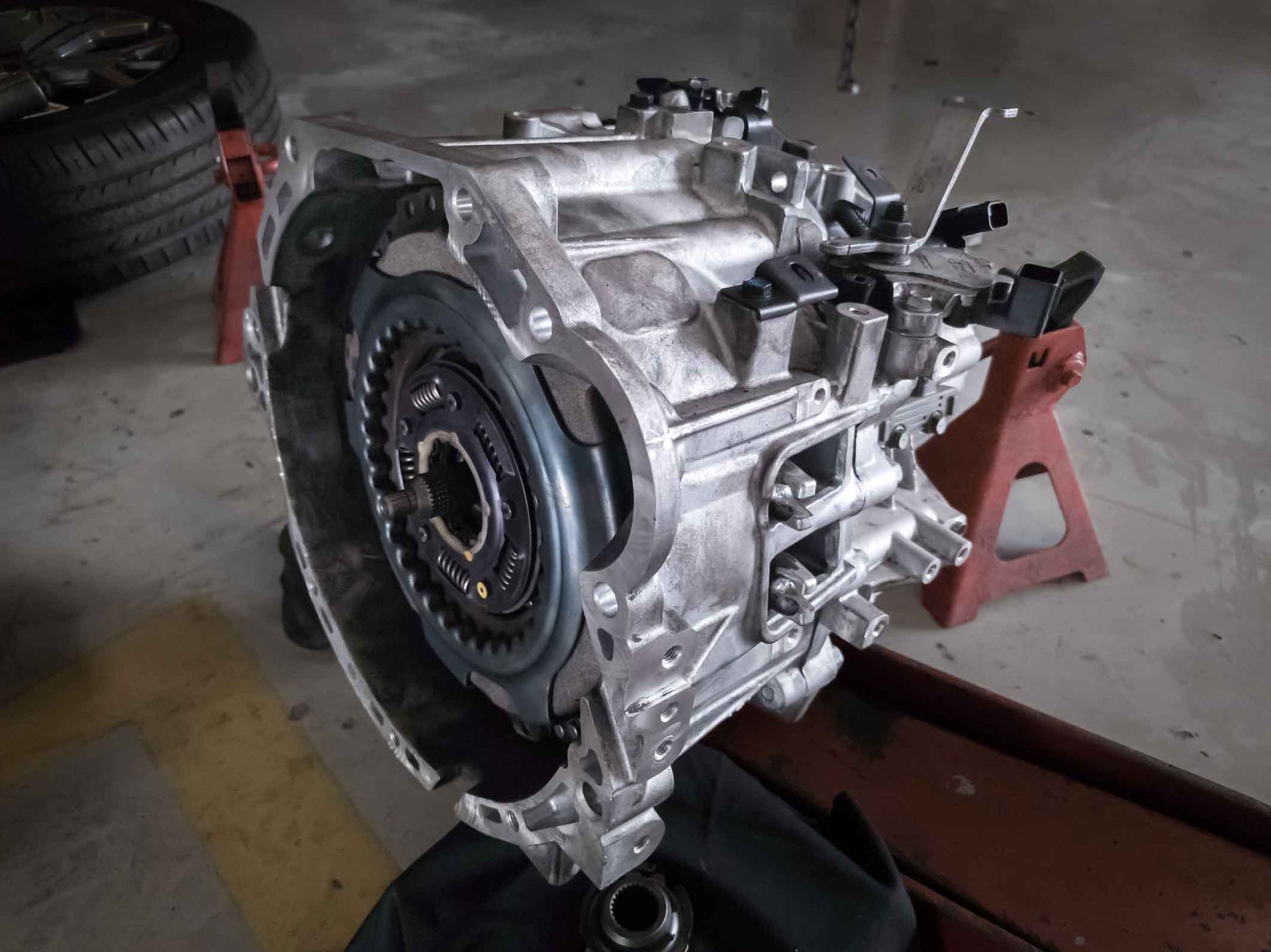 Car transmission assembly on a stand in a garage, showing the flywheel and metal casing.