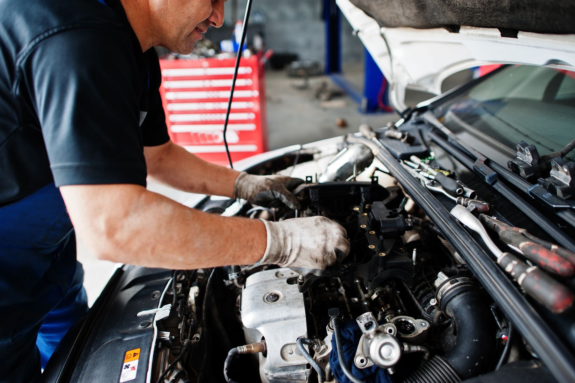 Mechanic working under a car hood in a garage, inspecting engine components with gloved hands.