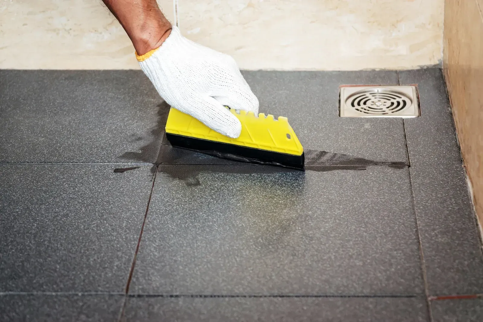 Gloved hand using a yellow tool to apply black sealant to tile grout in a shower.