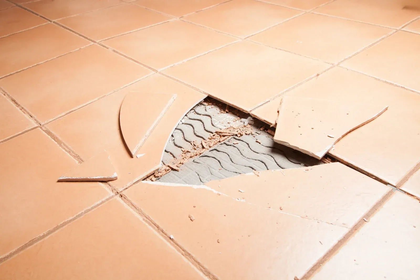Broken brown floor tiles showing the concrete underneath, with shattered tile pieces around the hole.