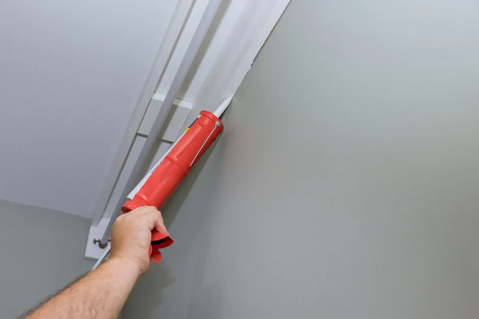 Hand applying caulk to a molding on a gray wall, using a red caulk gun.